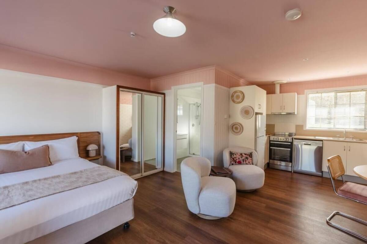 A bedroom with a bed, a bedside table, and a nightstand. There is a small seating area with a chair and ottoman. The room has a wooden floor and a pink wall.