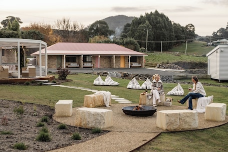 A covered outdoor seating area with stone seating and a fire pit, surrounded by a grassy landscape and a building with a red roof.