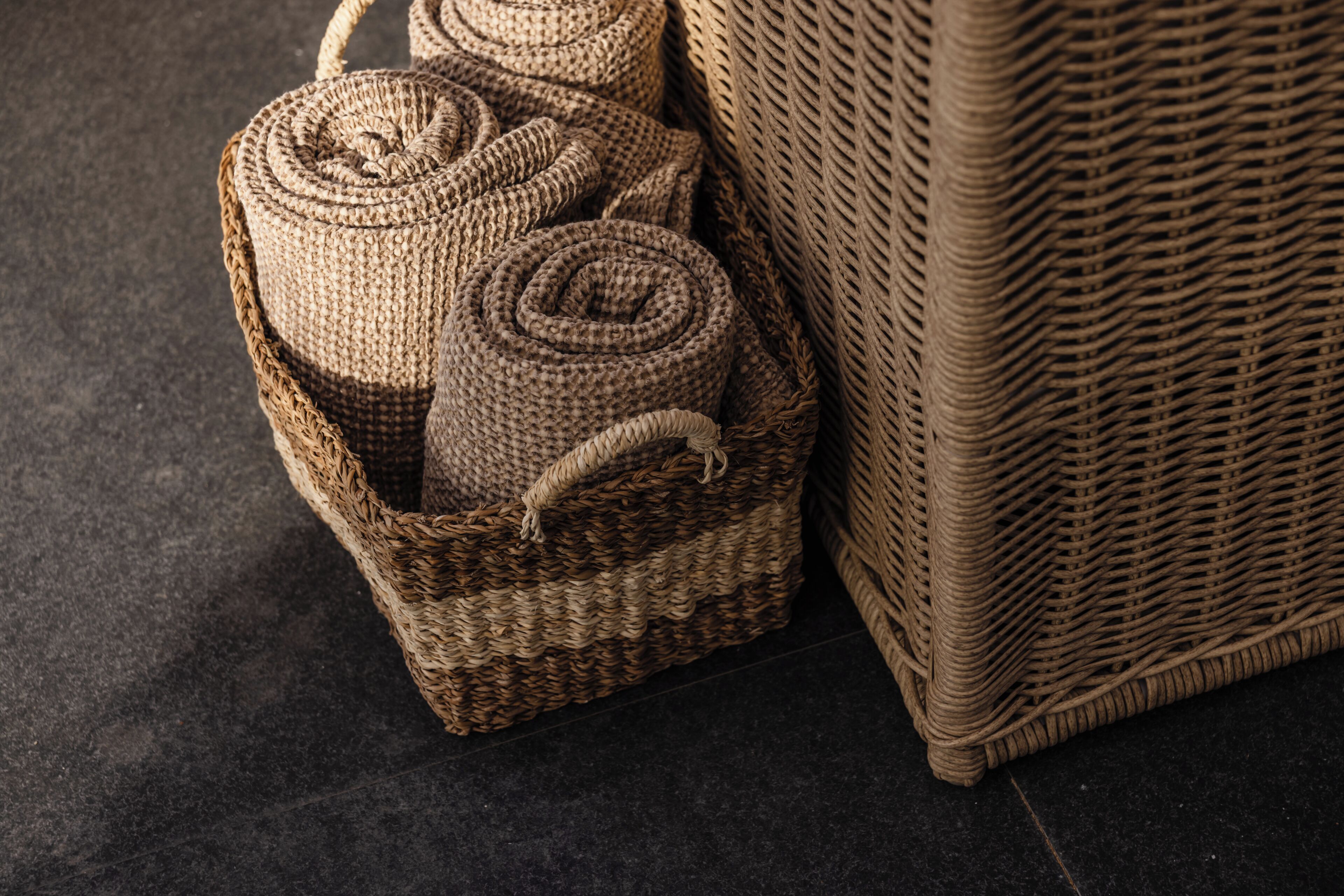 Una cesta de mimbre con objetos enrollados, junto a una silla de mimbre sobre un suelo oscuro.