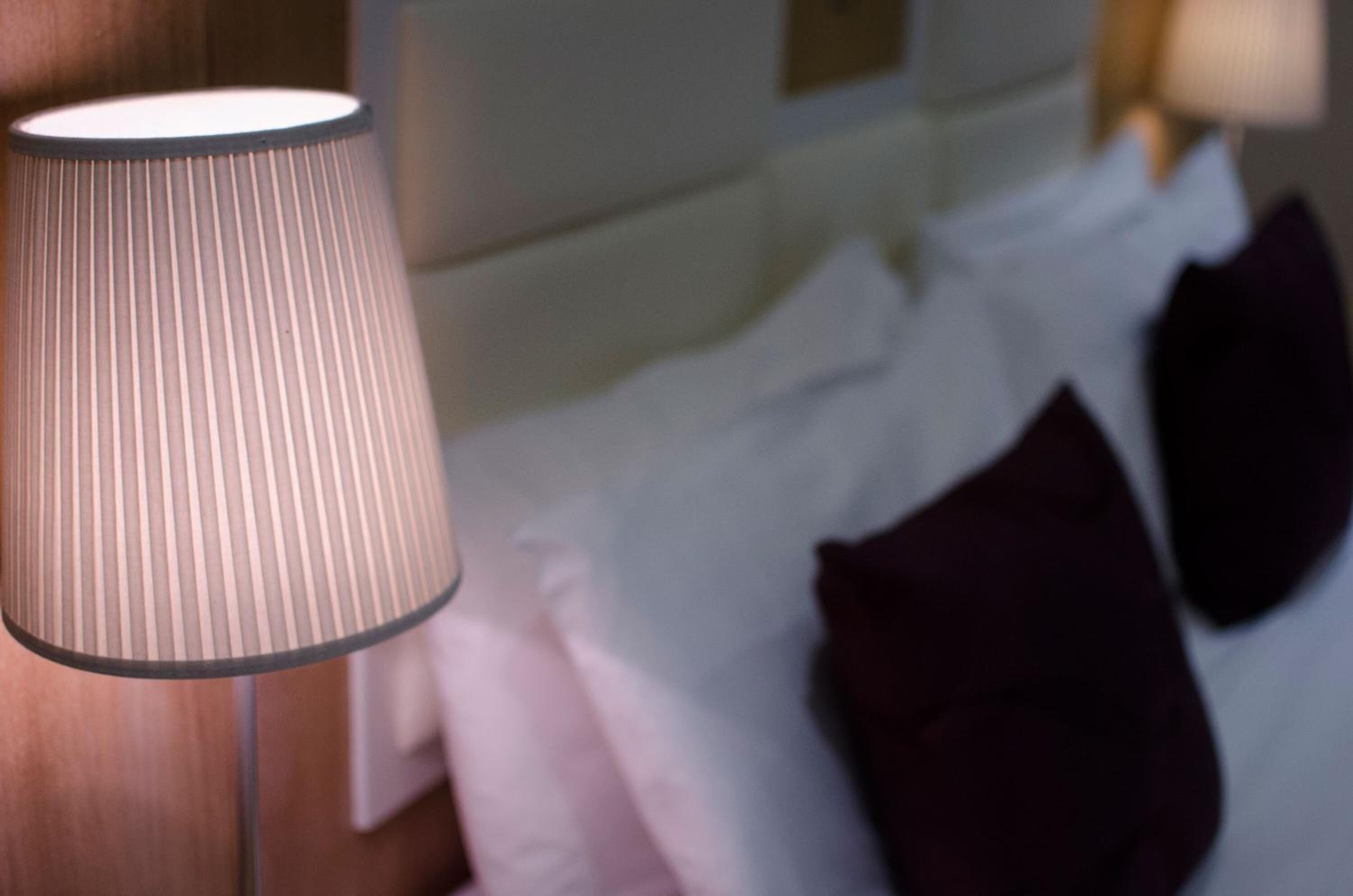 Eine Lampe mit geripptem Schirm, ein Bett mit weißer Bettwäsche und dunklen Kissen sowie ein Holzboden.