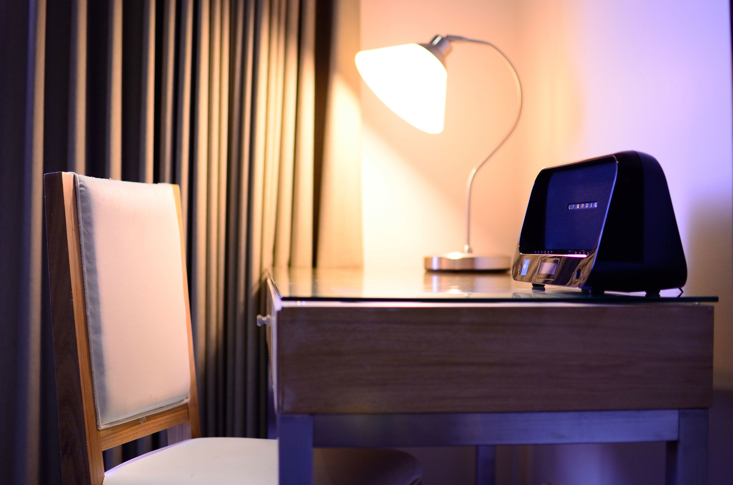 A hotel room with a wooden desk, a modern lamp, and a digital device on the desk.