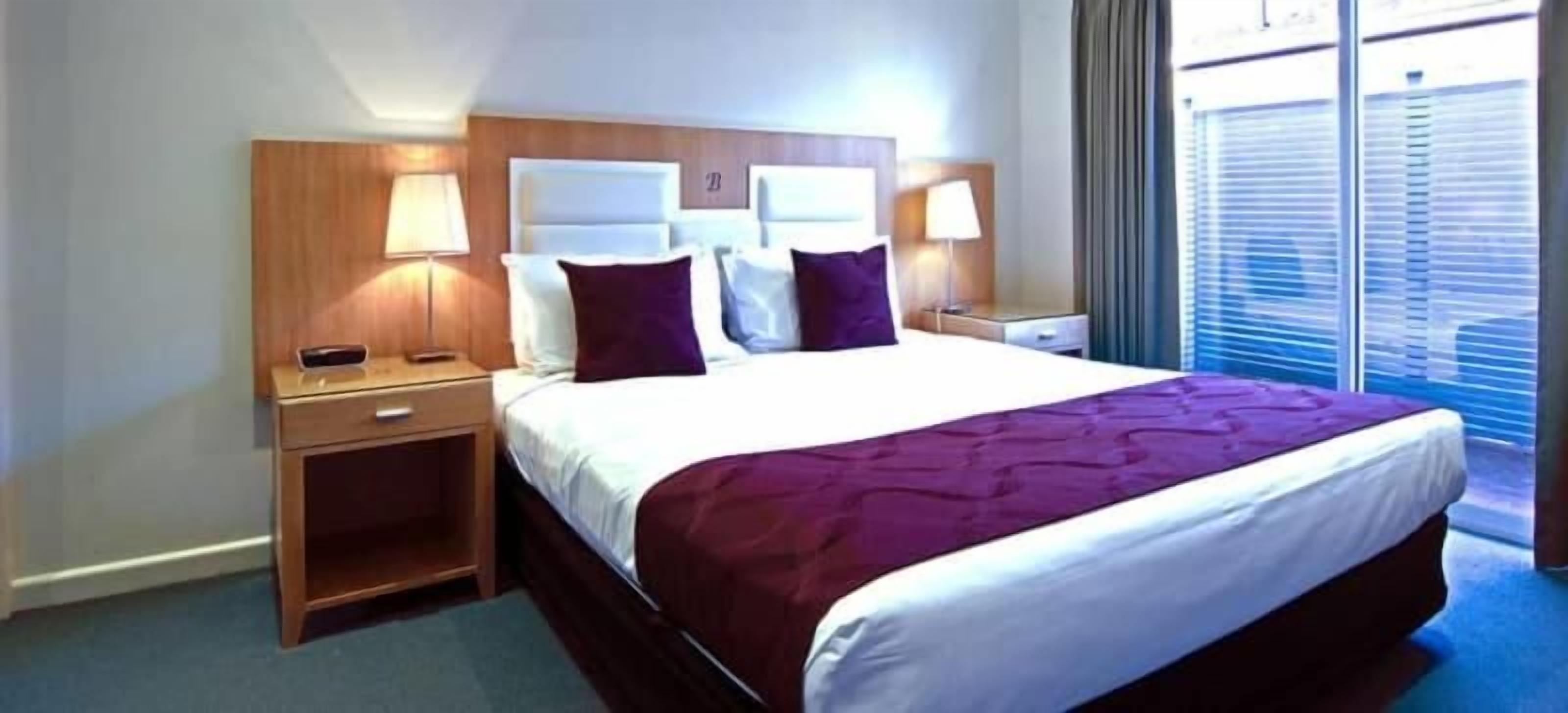 A hotel room with a large bed, two bedside tables, a nightstand, a window with blinds, and a view of the outside.