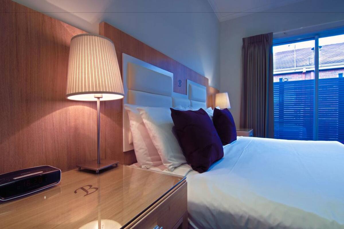 A hotel room with a bed, a bedside table with a lamp, a digital clock, and a window with curtains.