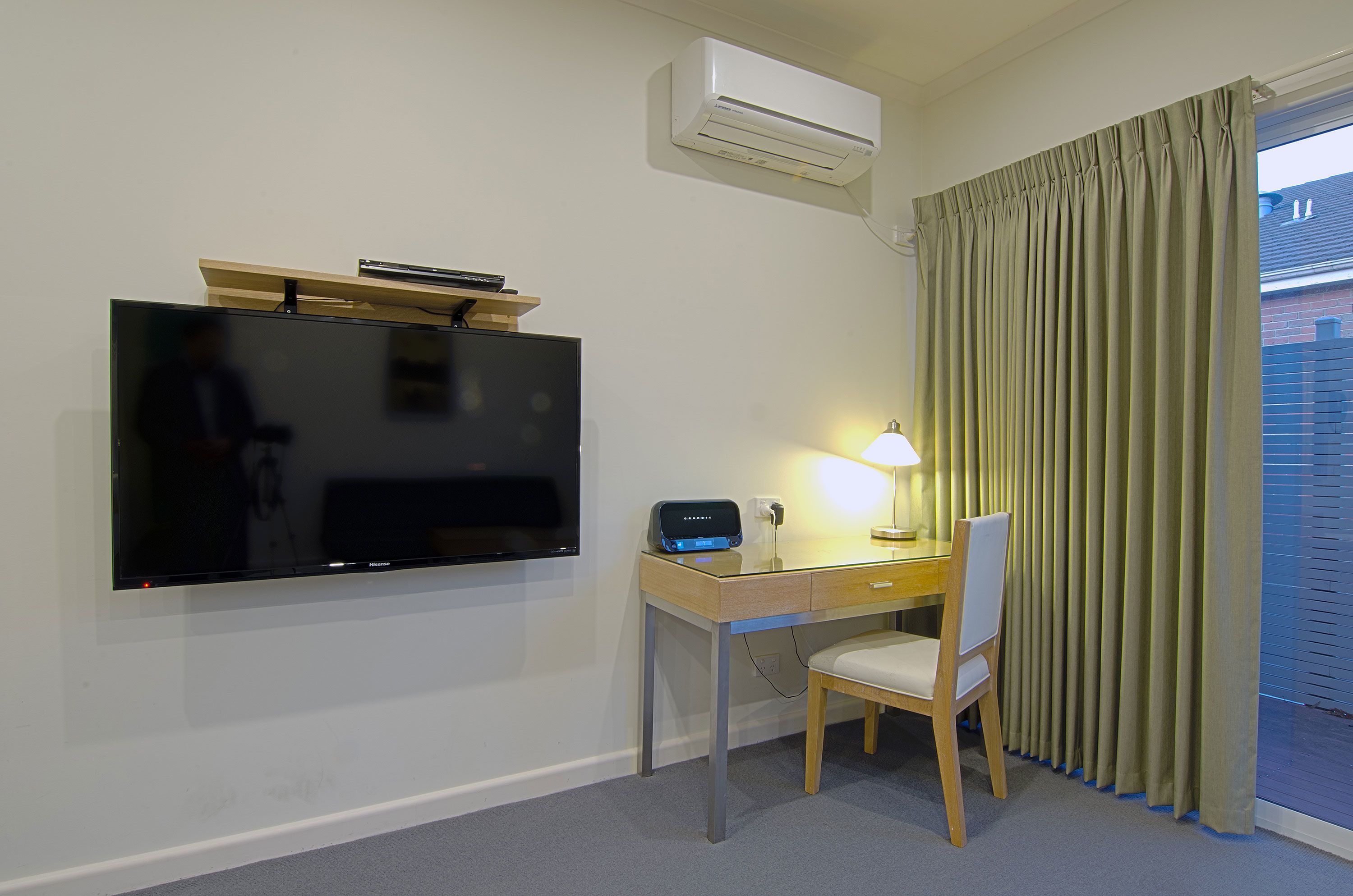 A hotel room with a flat-screen TV mounted on the wall, a desk with a lamp, a chair, and a window with curtains.