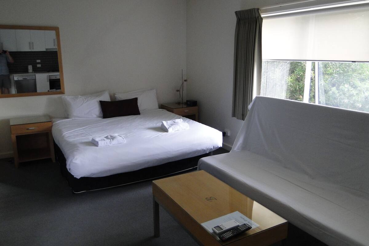 A hotel room with a bed, a sofa, a nightstand, a table with a remote control, and a window with curtains.
