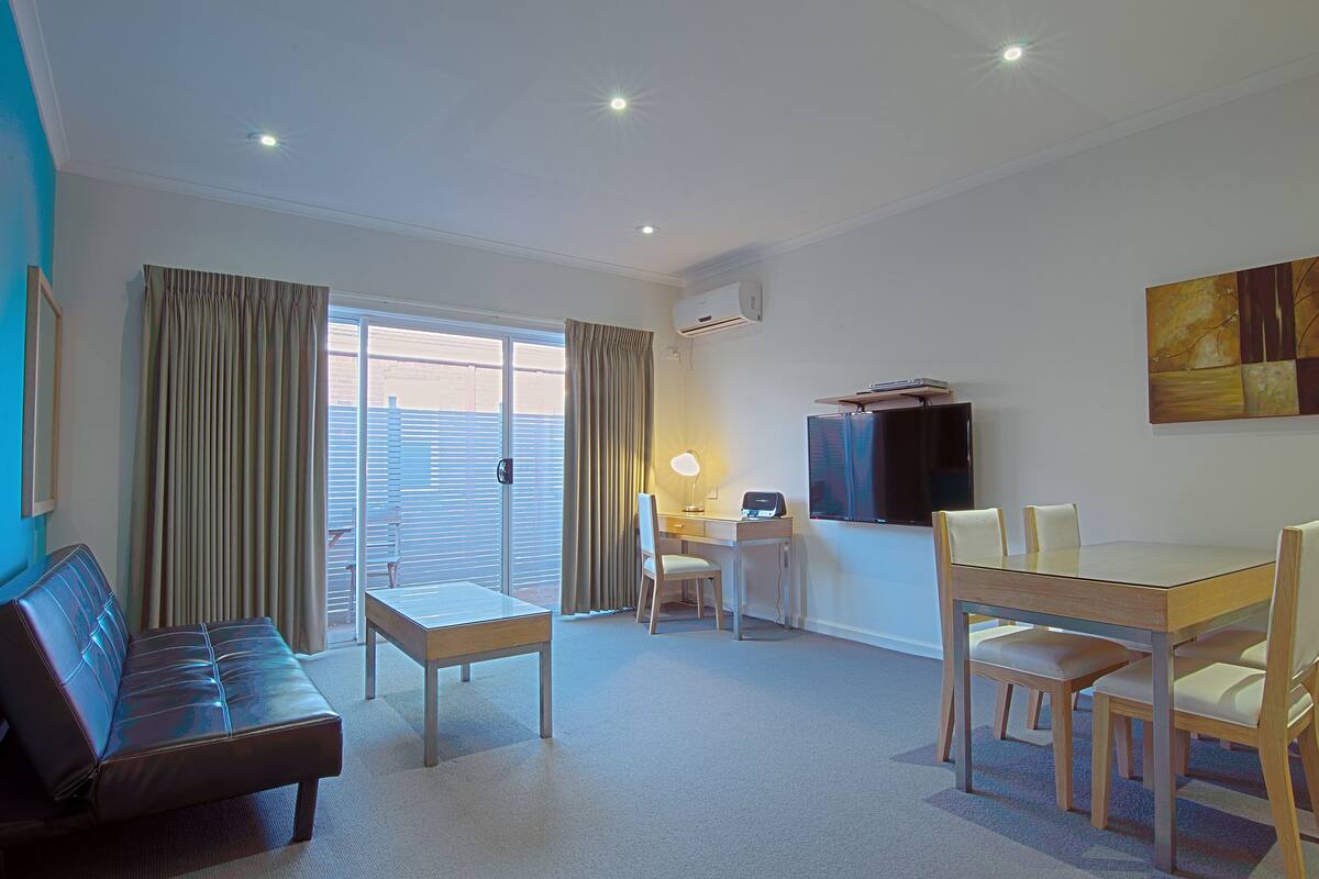 A hotel room with a sofa, a dining table with chairs, a desk with a chair, a TV mounted on the wall, and an air conditioning unit.
