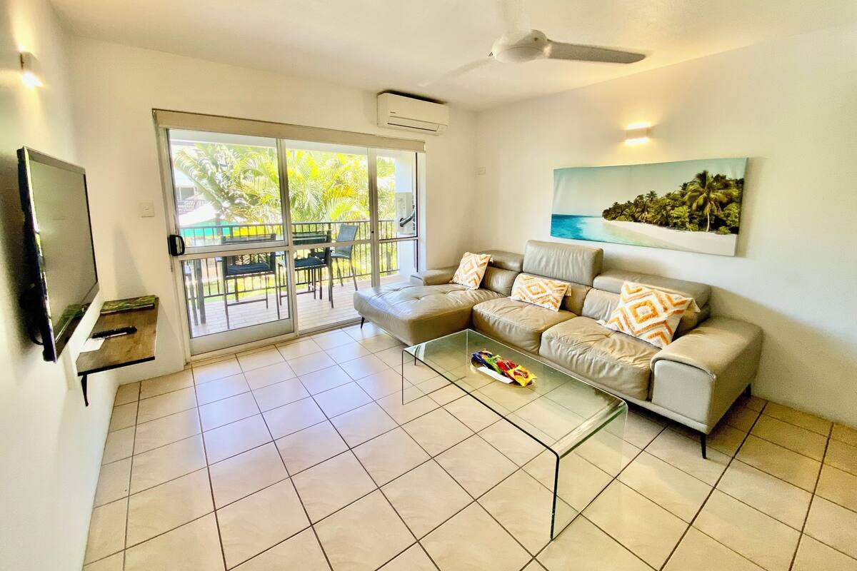 A living room with a sectional sofa, a glass coffee table, a flat-screen TV, and a balcony with a view of greenery.