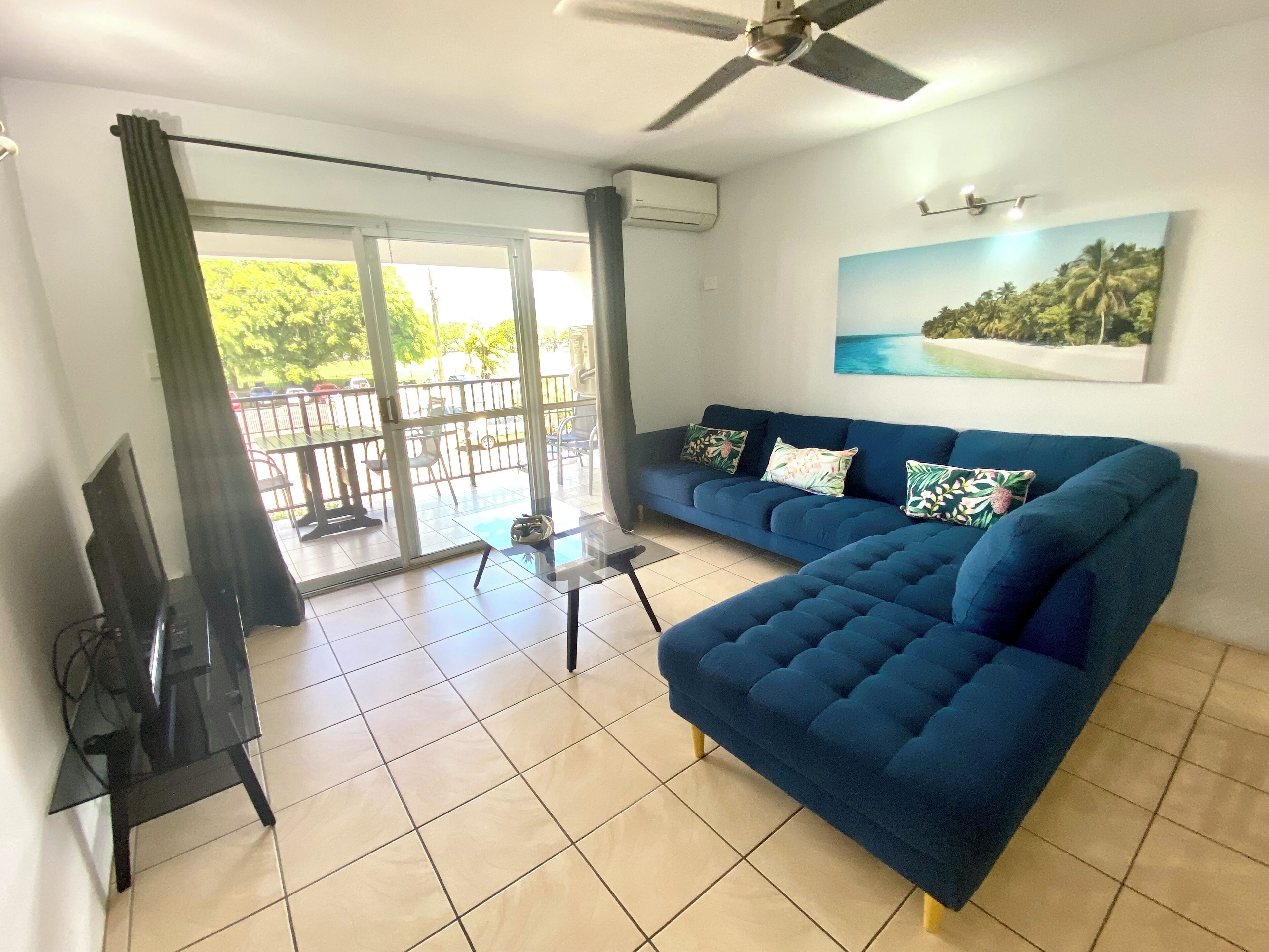 A living room with a blue sofa, a glass coffee table, a TV, and a balcony with a view of greenery.