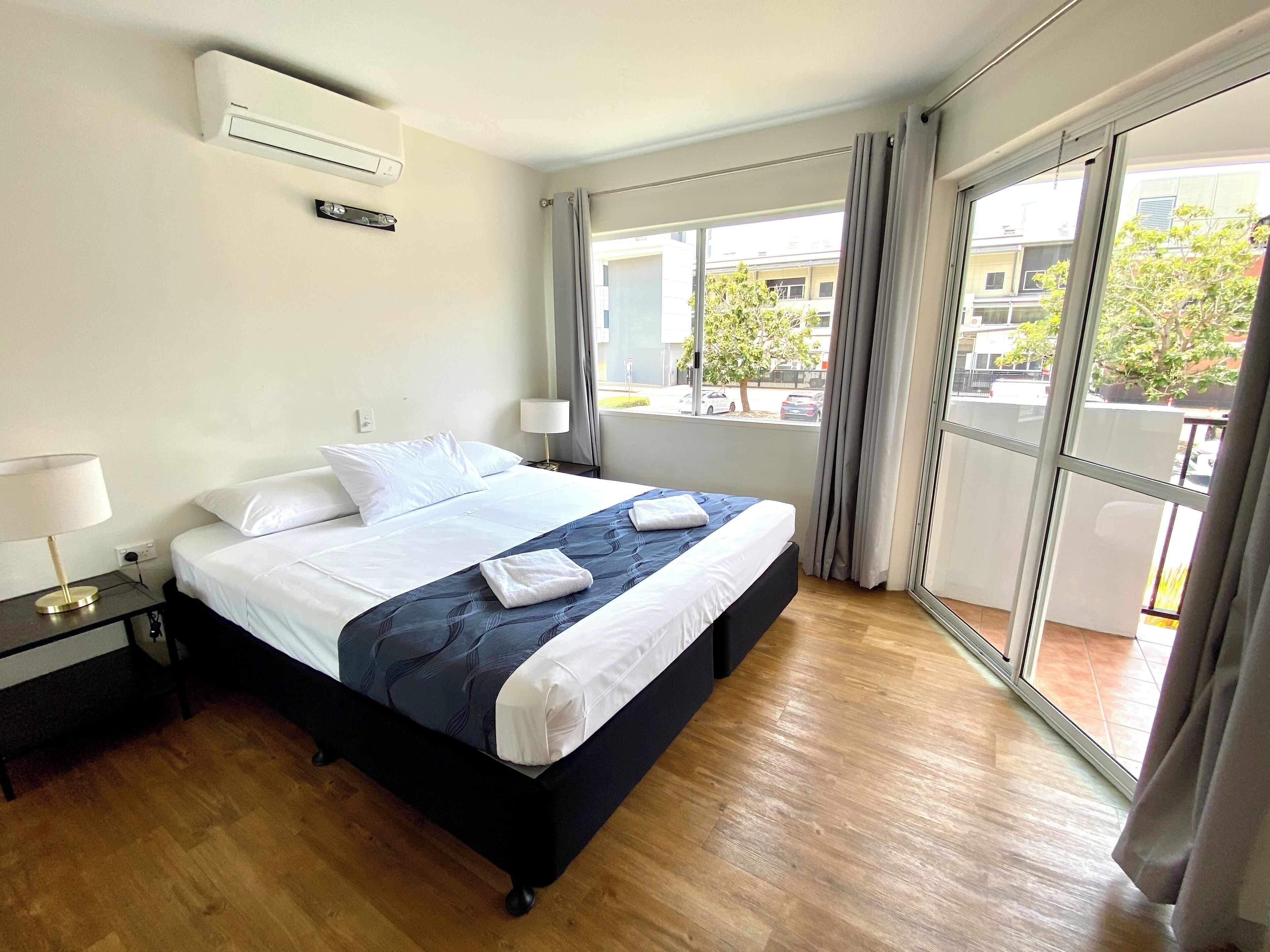 A hotel room with a bed, a nightstand, a lamp, an air conditioning unit, and a view of the outside through a large window.