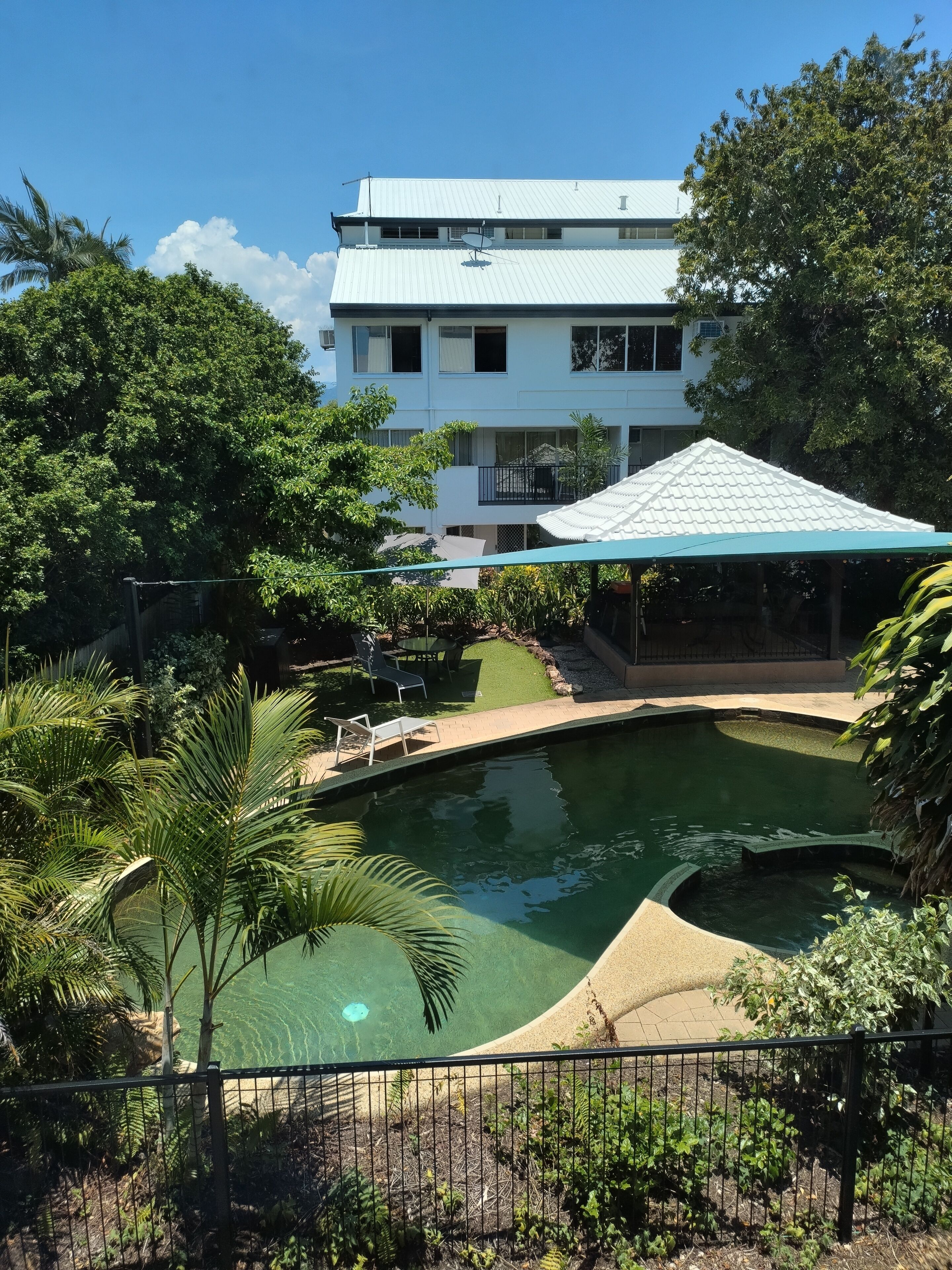 A swimming pool surrounded by lush greenery, a covered seating area, and a multi-story building in the background.