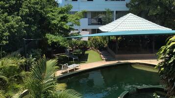 A swimming pool surrounded by lush greenery, a covered seating area, and a multi-story building in the background.