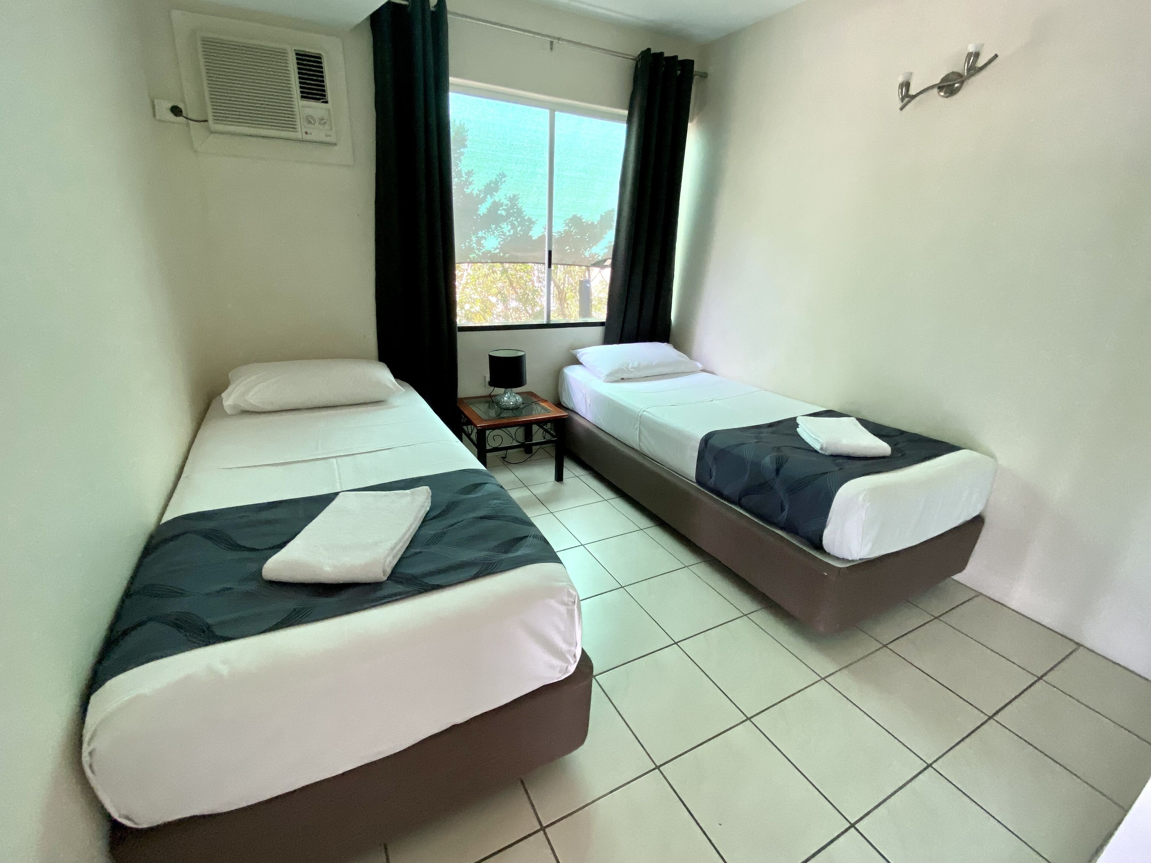 Quarto com duas camas de solteiro, uma mesinha com abajur e uma janela com cortinas.