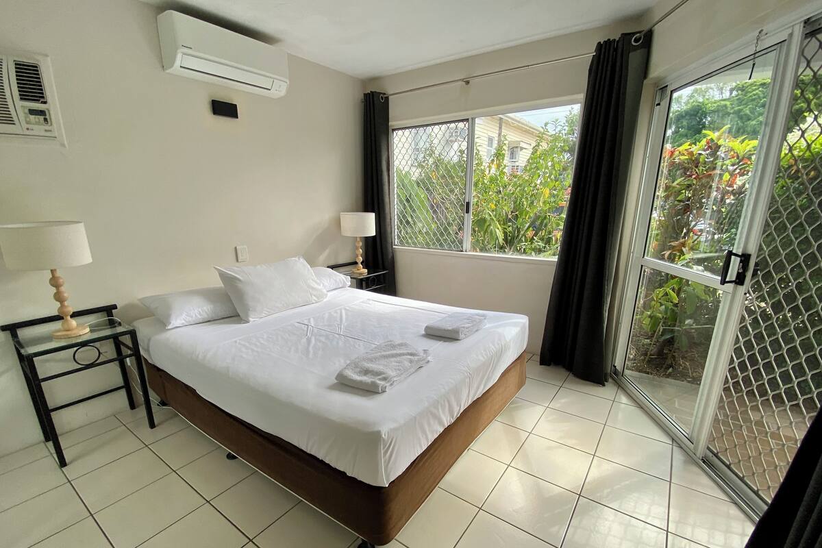 A hotel room with a bed, two bedside tables, a lamp, an air conditioner, and a sliding glass door to a balcony.