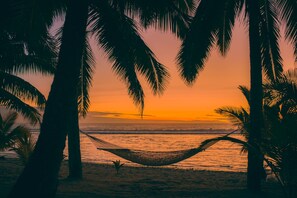 A hammock on a beach with palm trees and a sunset.