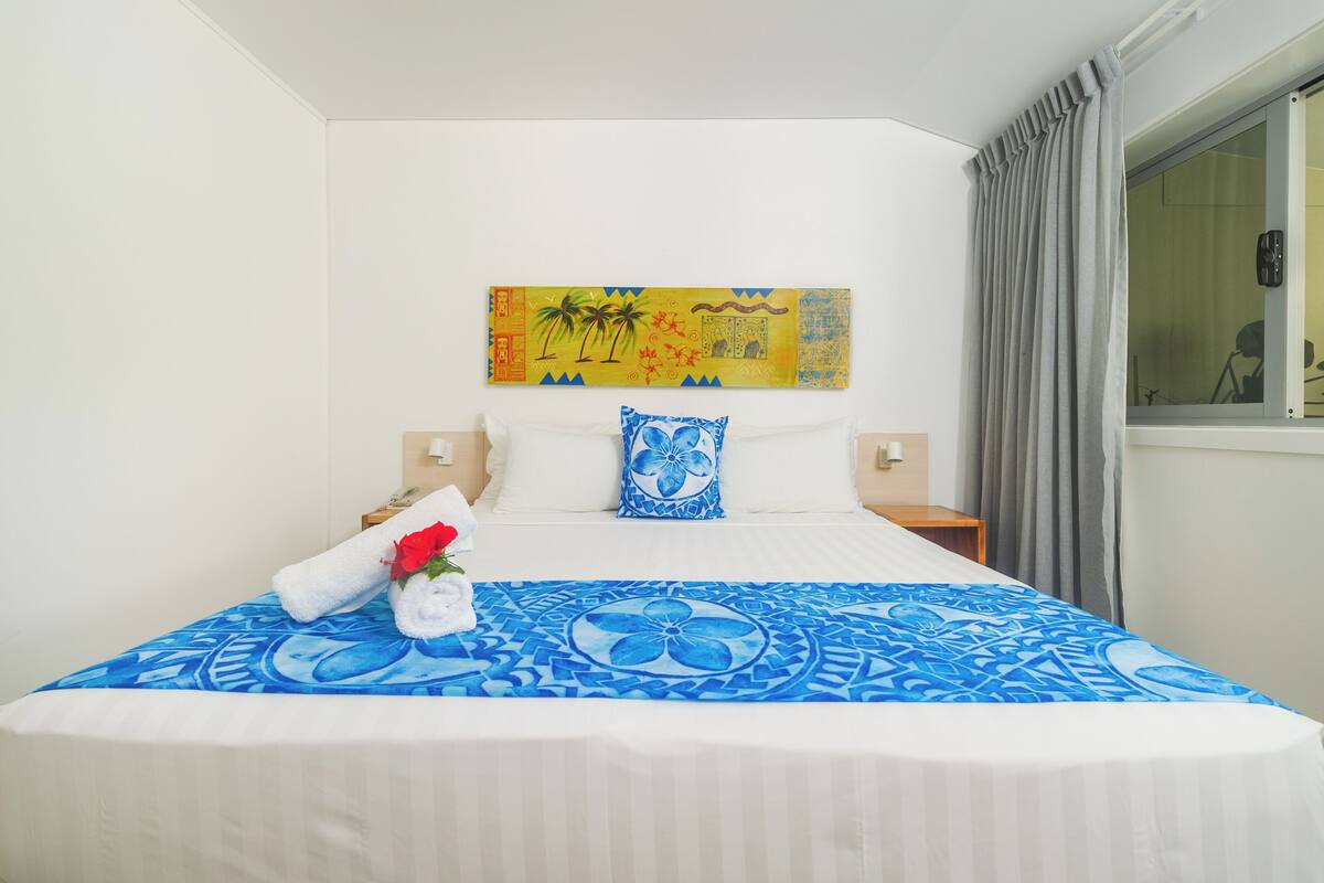 A hotel room with a bed, a painting on the wall, a window with curtains, and a nightstand with a lamp.