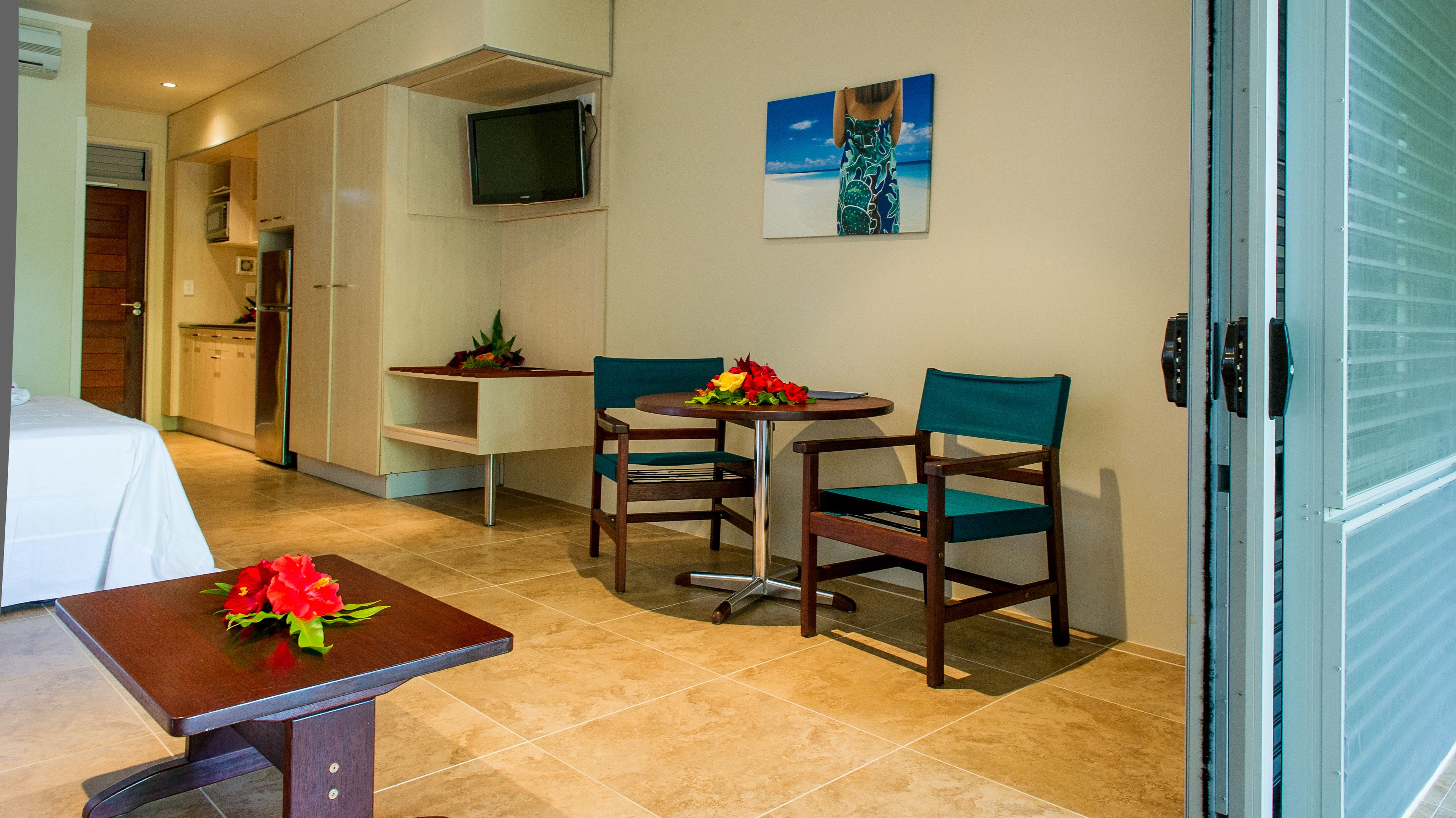 A hotel room with a bed, a small table with a vase of flowers, two chairs, a television mounted on the wall, and a painting above the chairs.