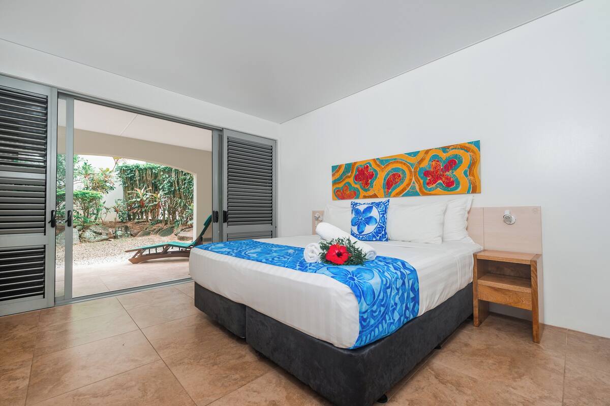 A bedroom with a bed, a nightstand, a painting, and a view of an outdoor area with a table and chairs.