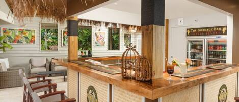 Zona de bar con mobiliario de madera, una jaula de pájaros y vistas al exterior del complejo turístico.