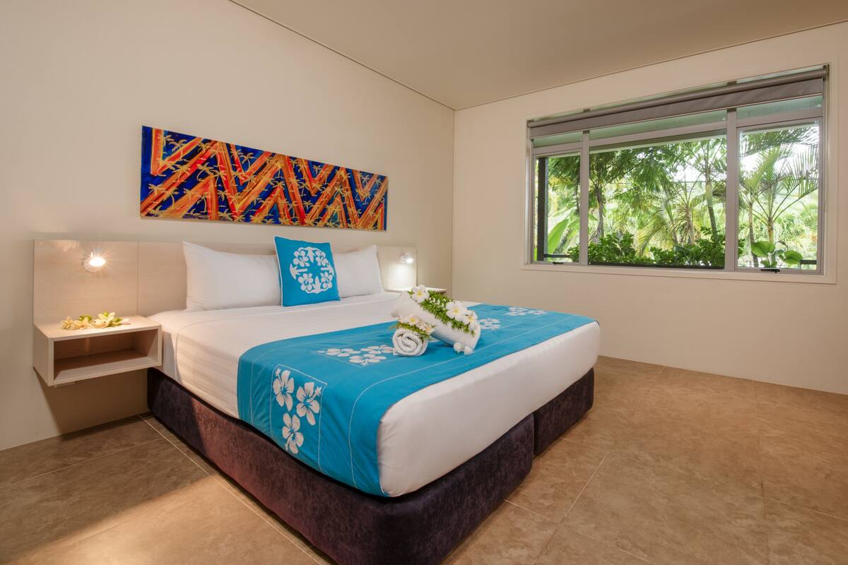 A modern hotel room with a large bed, a nightstand, a window with a view of trees, and a colorful abstract painting on the wall.