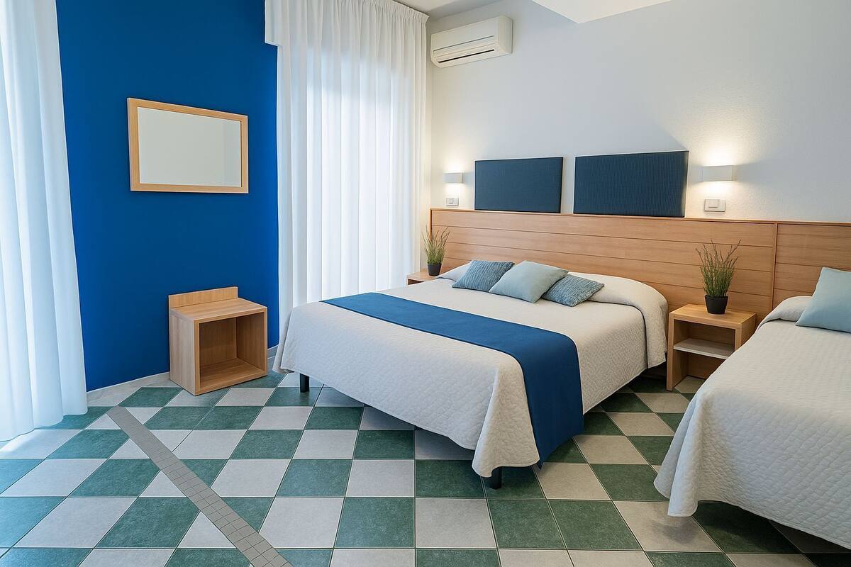 A hotel room with a checkered floor, a large bed with a blue accent, a bedside table with a lamp, and a wall-mounted air conditioner.