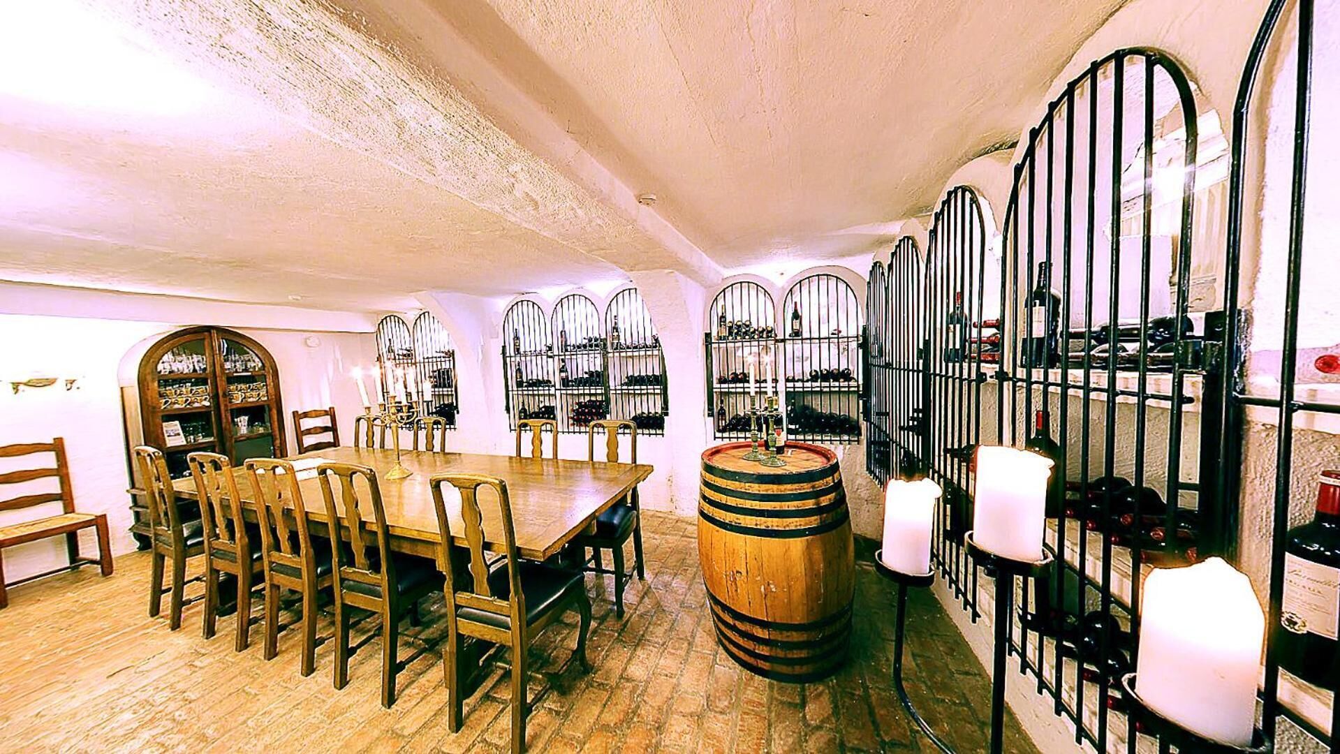 Comedor con mesa y sillas de madera, un barril de vino y una botellera con botellas.