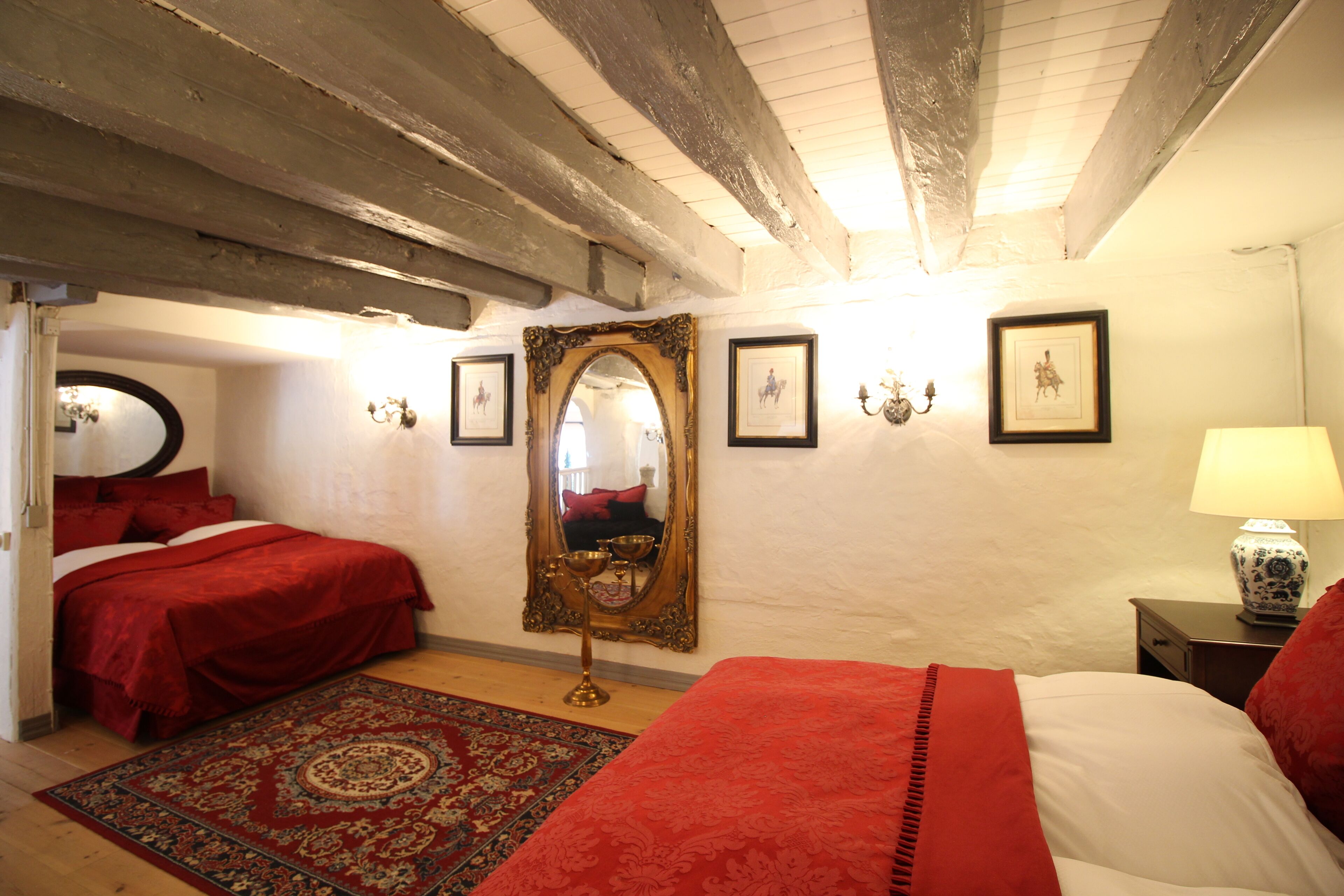 Een slaapkamer met een groot bed, een kleiner bed, een spiegel, ingelijste foto's, een lamp en een rood vloerkleed.