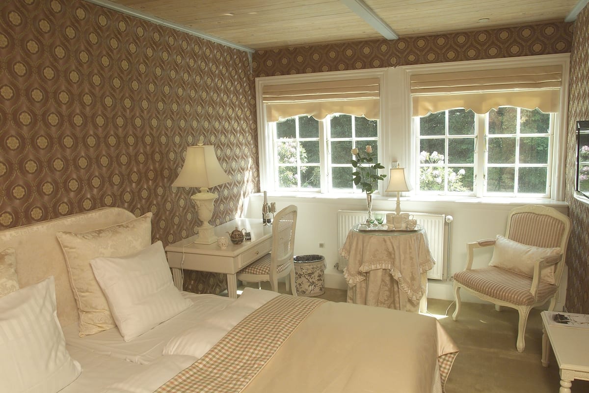 A bedroom with a large bed, a desk, a chair, and a window with curtains.