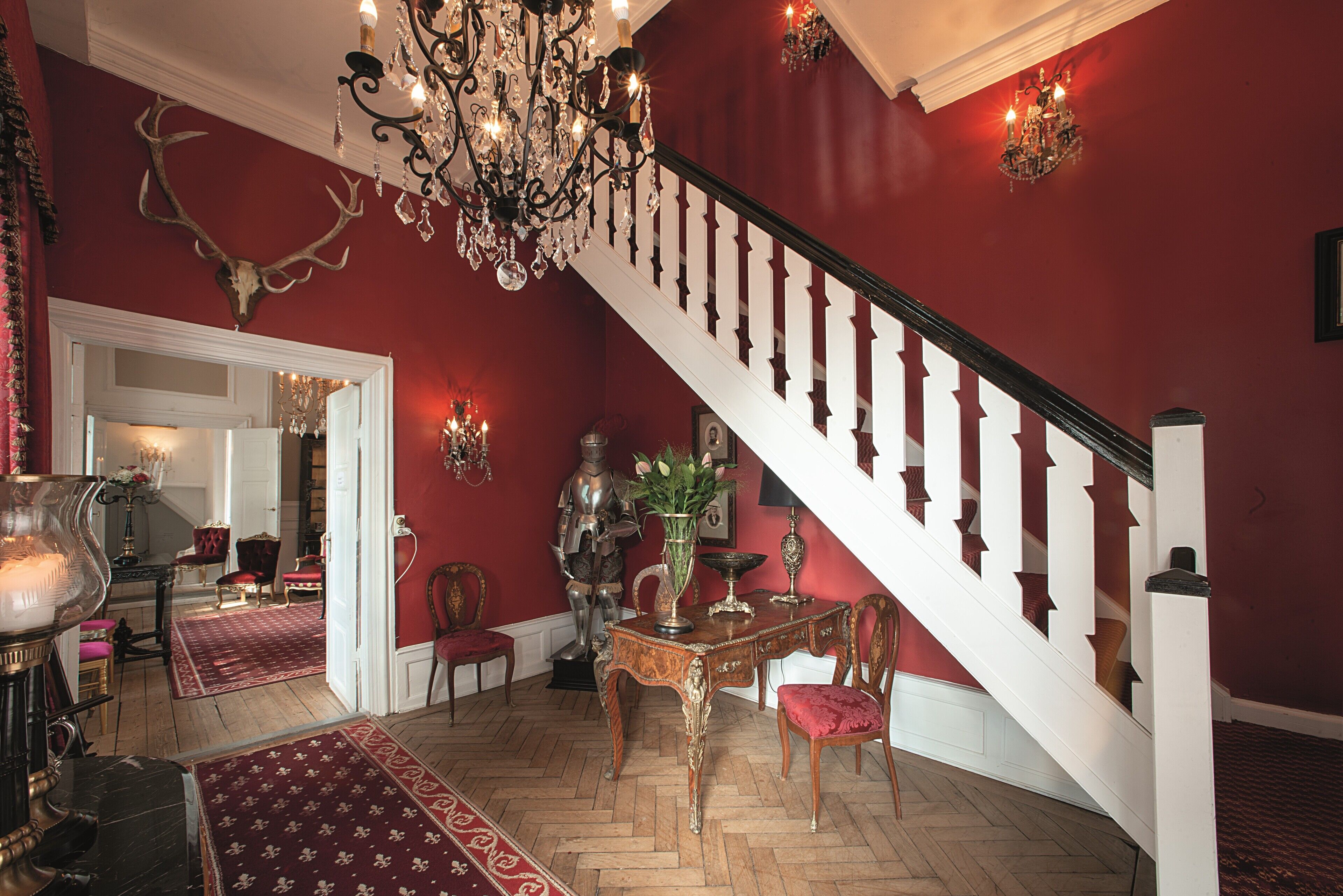 A grand staircase with white balusters and a red wall. A chandelier hangs from the ceiling, and a deer skull decorates the wall.