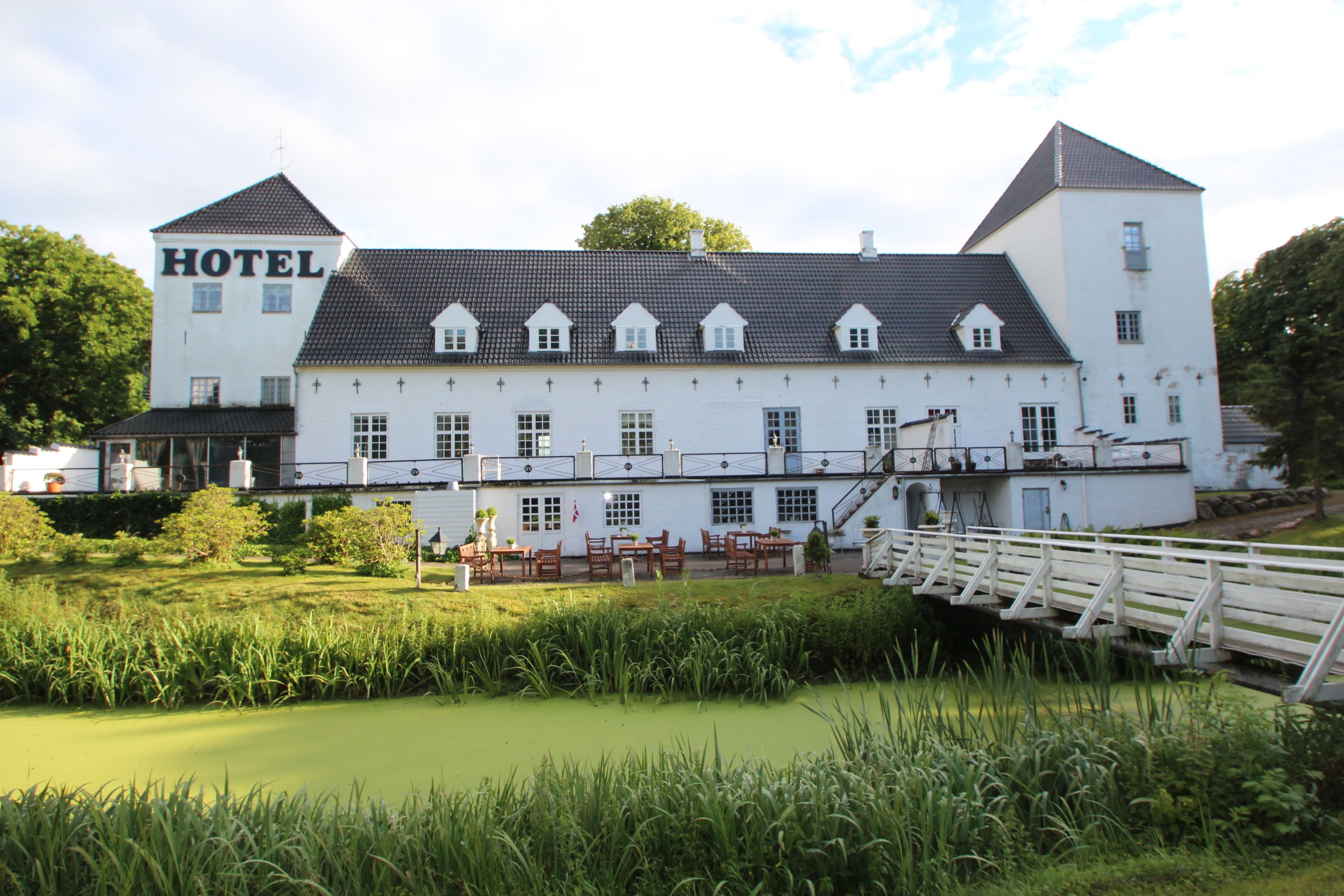 A large white building with a sign reading "HOTEL" on the side. It has multiple windows, a covered outdoor seating area, and a small bridge over a pond.
