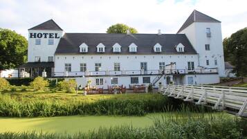 A large white building with a sign reading "HOTEL" on the side. It has multiple windows, a covered outdoor seating area, and a small bridge over a pond.
