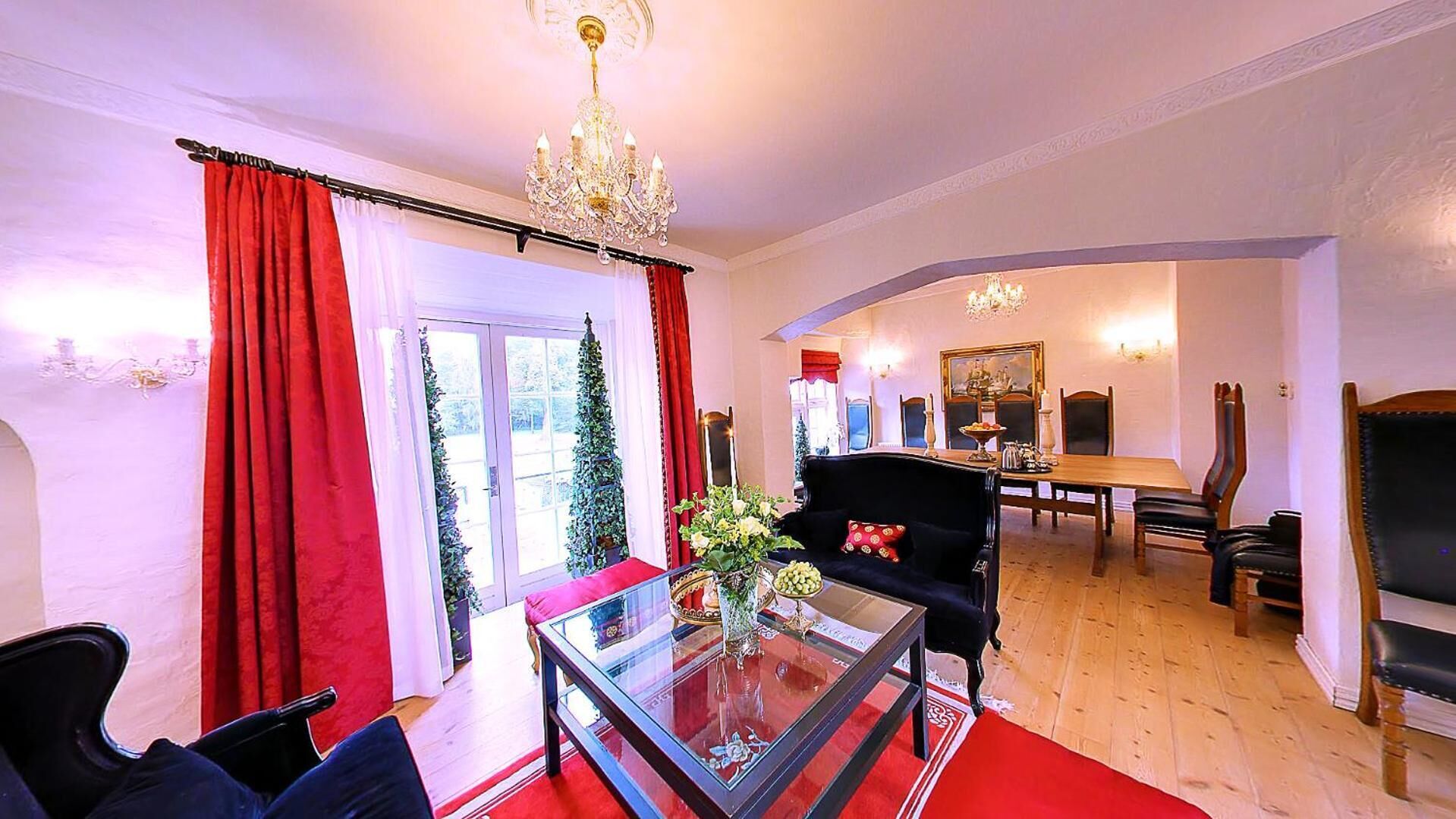 A spacious living room with a glass coffee table, red curtains, a chandelier, and a dining area with a painting on the wall.