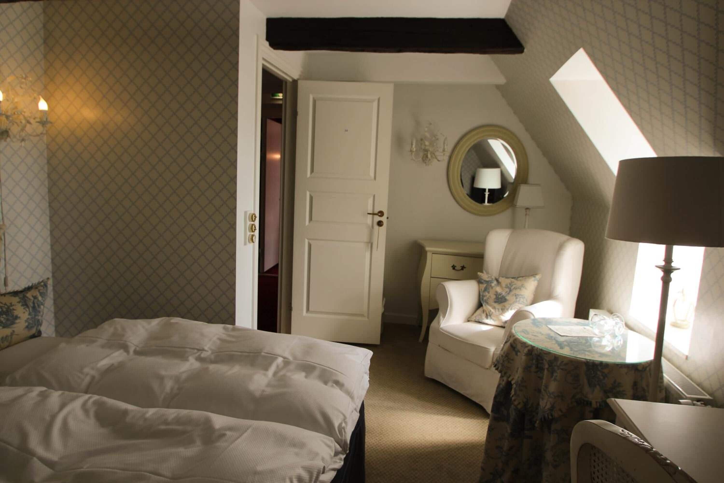 A bedroom with a bed, a chair, a round table, a mirror, and a door.