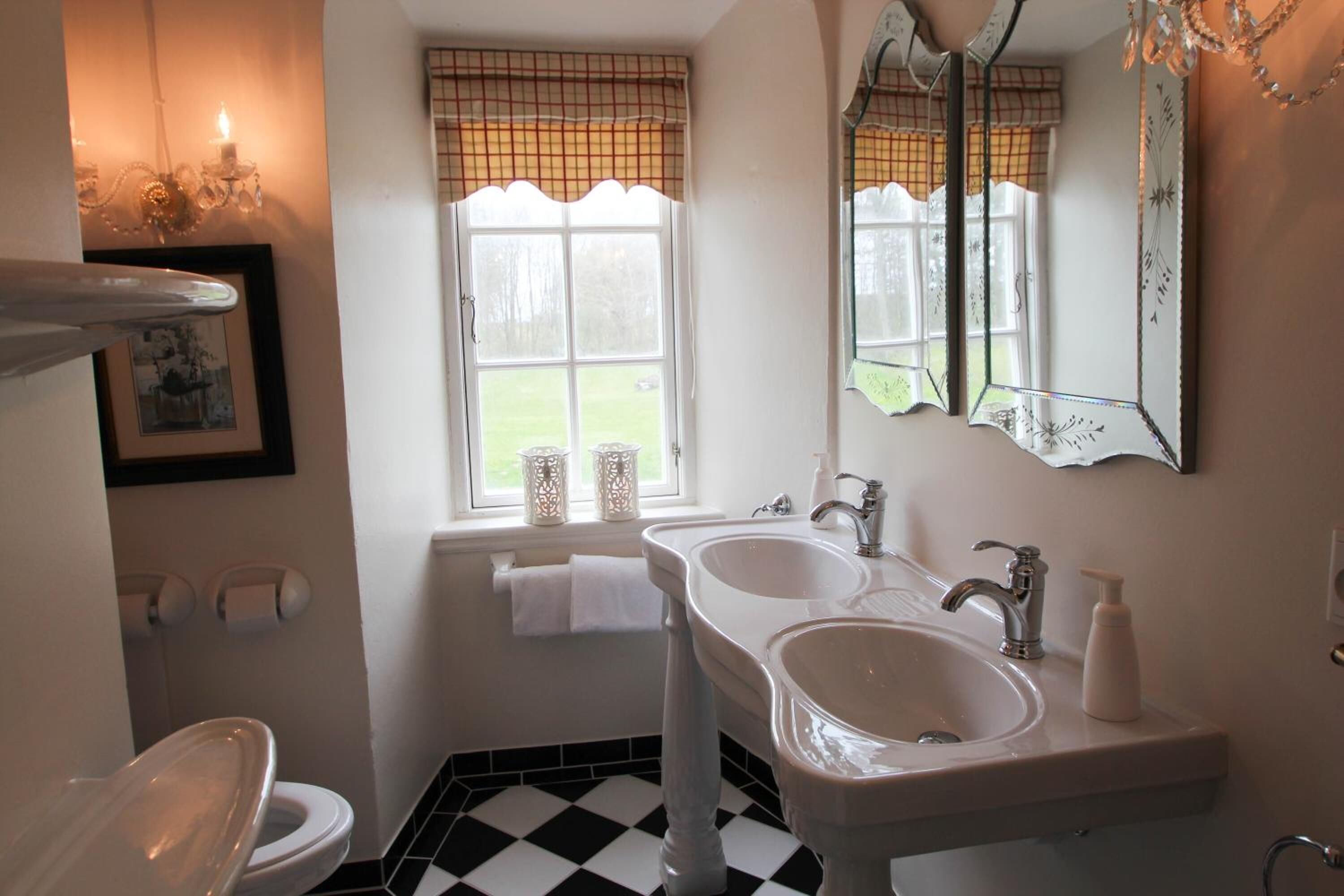 Un baño con dos lavamanos, inodoro, ventana con cortina de cuadros y candelabro.