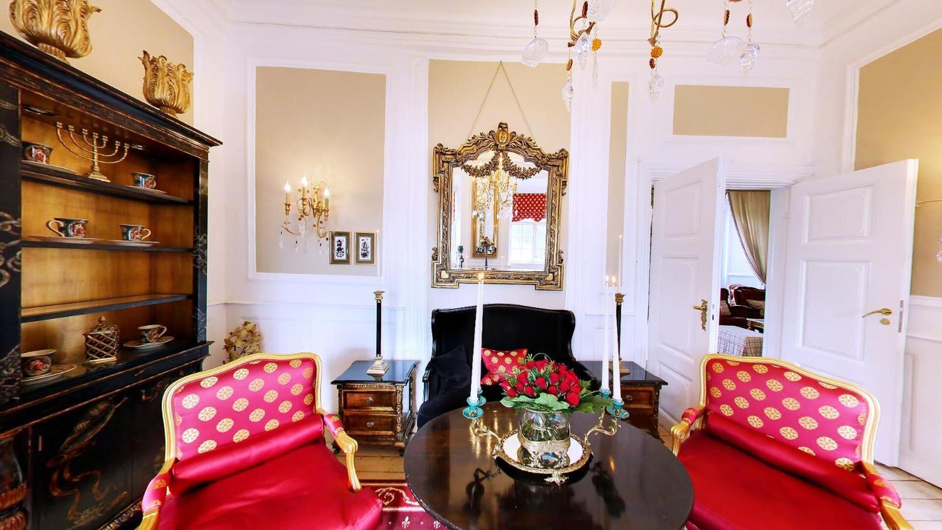 Une salle à manger élégante, avec une table noire, des chaises à l’assise rouge, un lustre et un grand miroir décoratif.