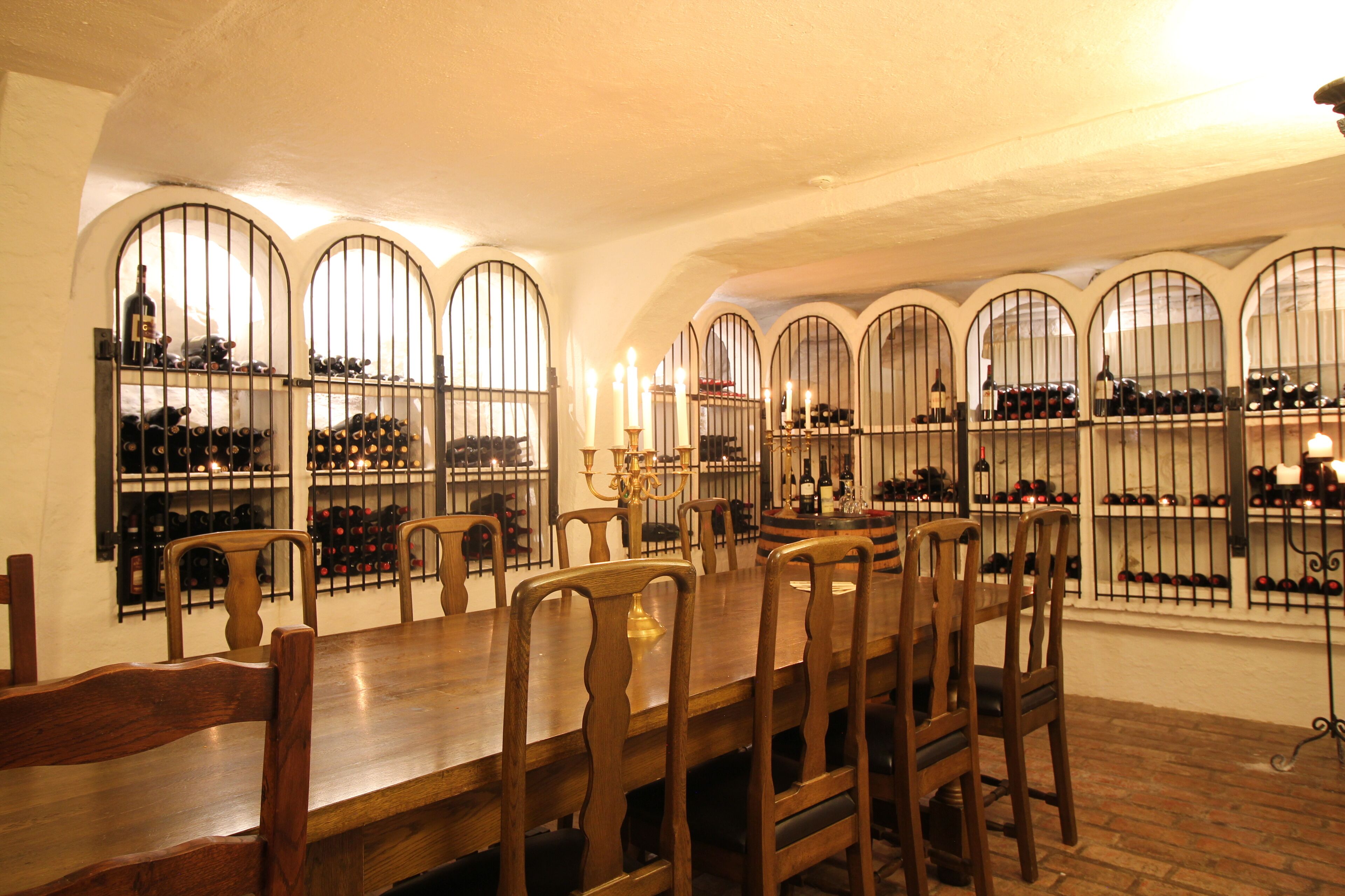 Un espace repas avec une longue table en bois et des chaises, entouré de fenêtres en arc et de présentoirs à bouteilles de vin.
