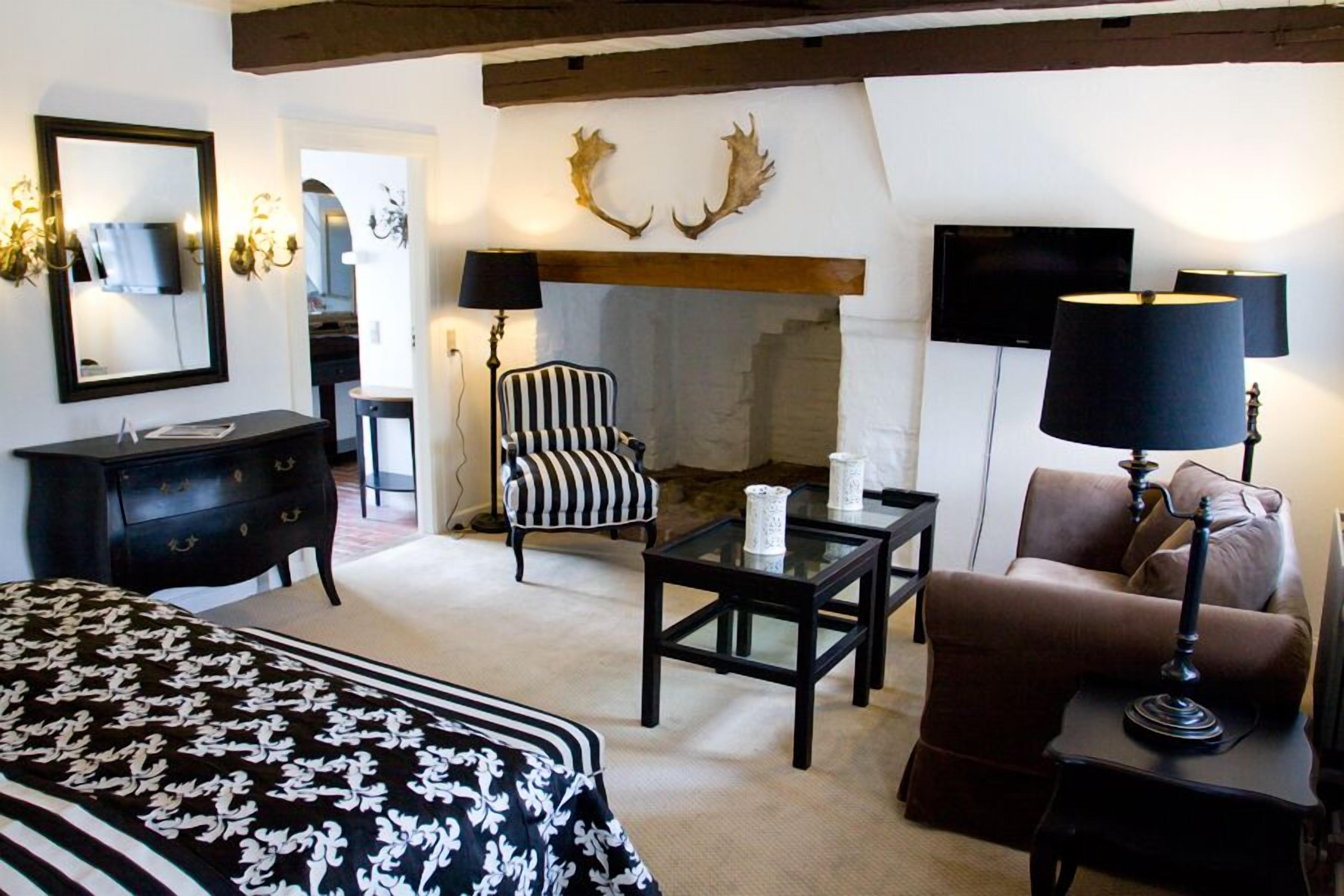 Una habitación de hotel con cama, chimenea, sillón a rayas, sofá marrón, lámpara negra y televisor.