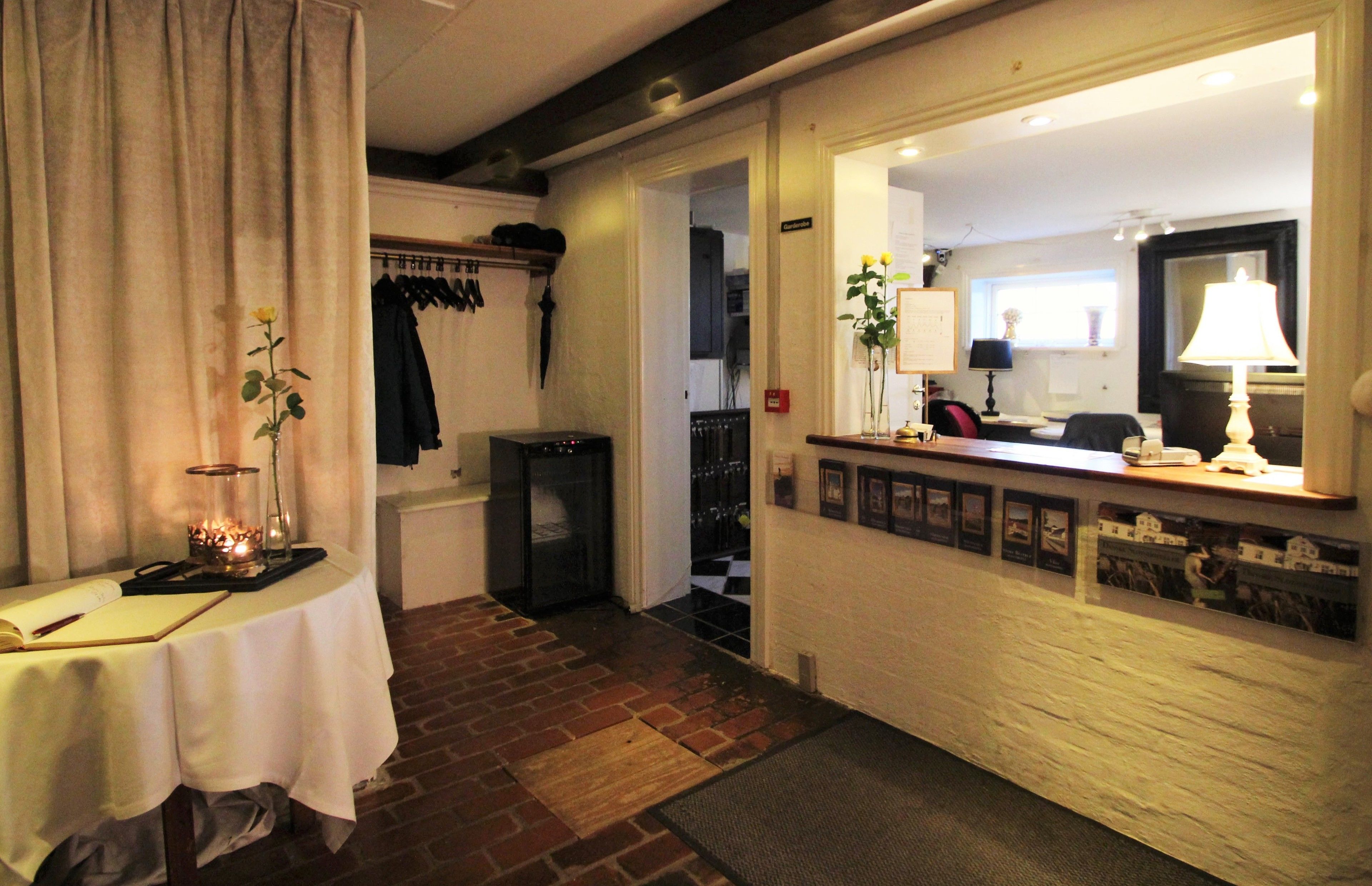 A hotel room with a brick floor, a table with a book and a candle, a coat rack, and a door leading to another room.