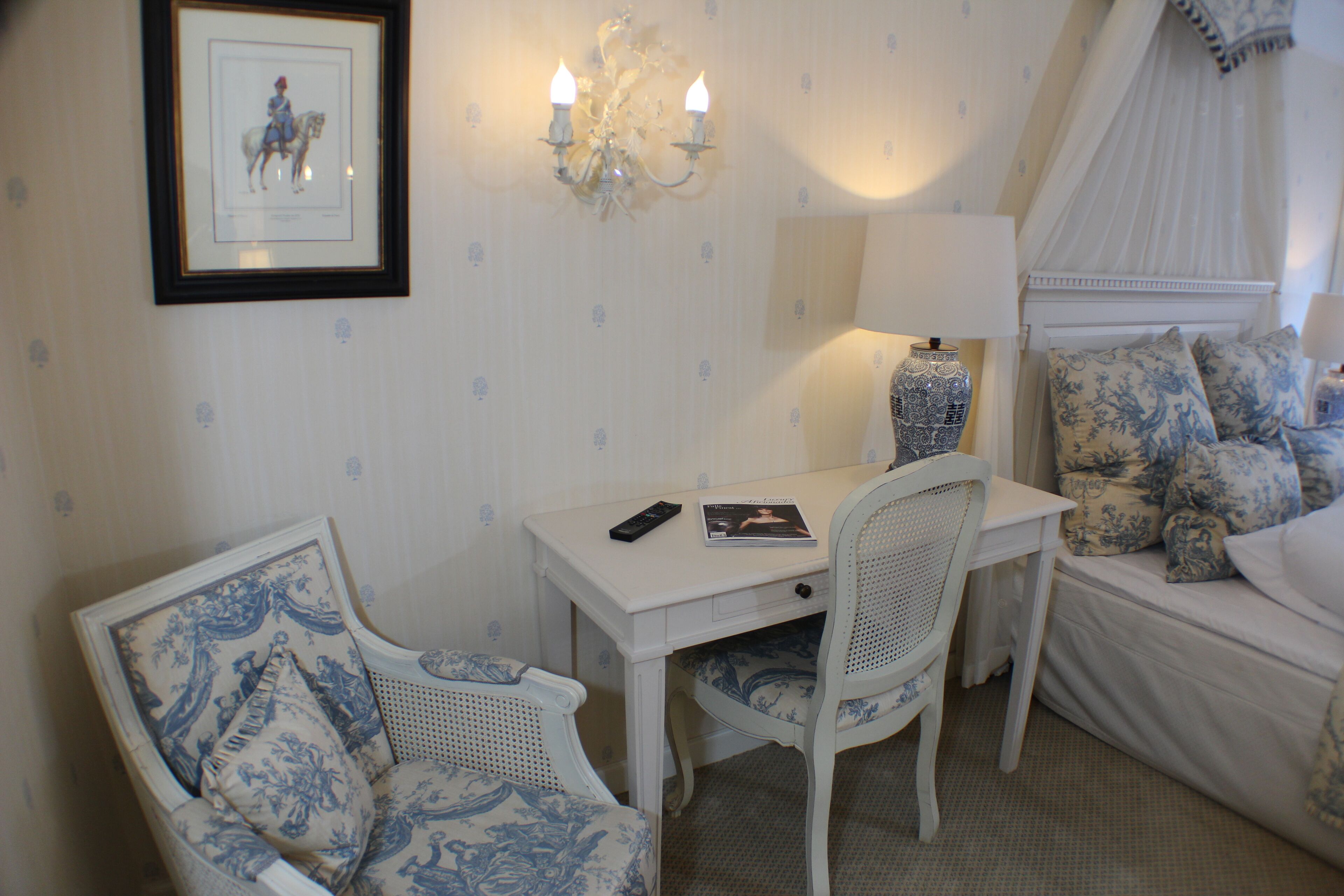 A room with a white bed, a white desk, a white chair, a chandelier, and a framed picture on the wall.