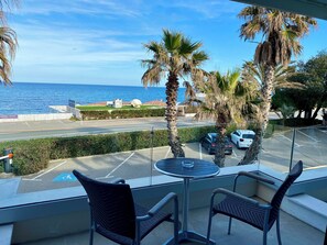 A balcony with two chairs and a table overlooking a parking area, palm trees, and a view of the sea. - Hôtel La Plage (Sainte-Maxime)
