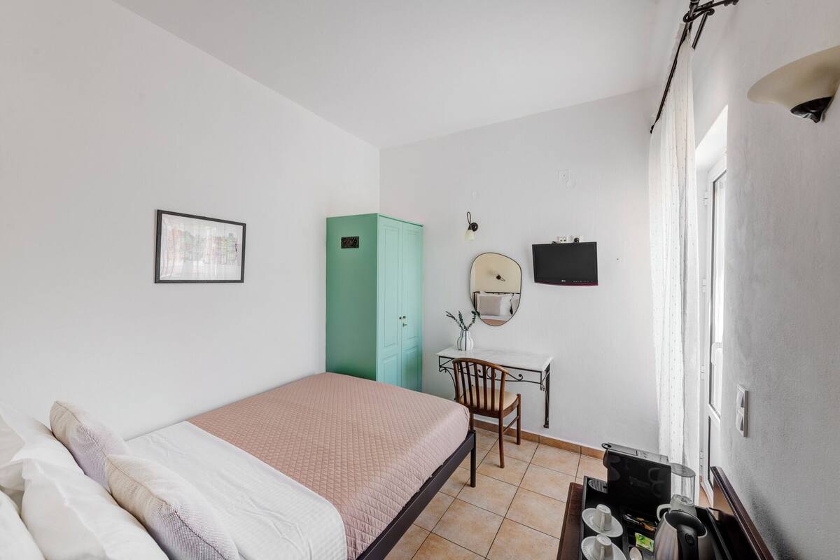A bedroom with a bed, a desk with a chair, a green wardrobe, a wall-mounted TV, and a framed picture.