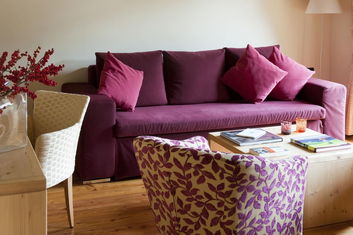 A cozy living room with a wooden ceiling, a purple sofa, a patterned armchair, and a small table with books and a vase of red flowers.