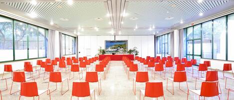 A conference room with rows of red chairs, large windows, and a ceiling with linear lighting fixtures.