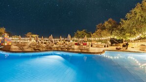 Piscina con sillas de descanso y sombrillas, rodeada de árboles y luces colgantes.