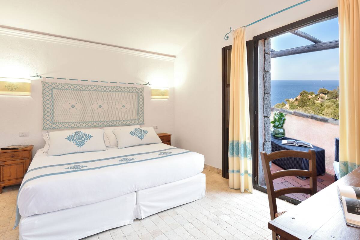 A bedroom with a bed, bedside tables, and a view of the sea through a glass door.