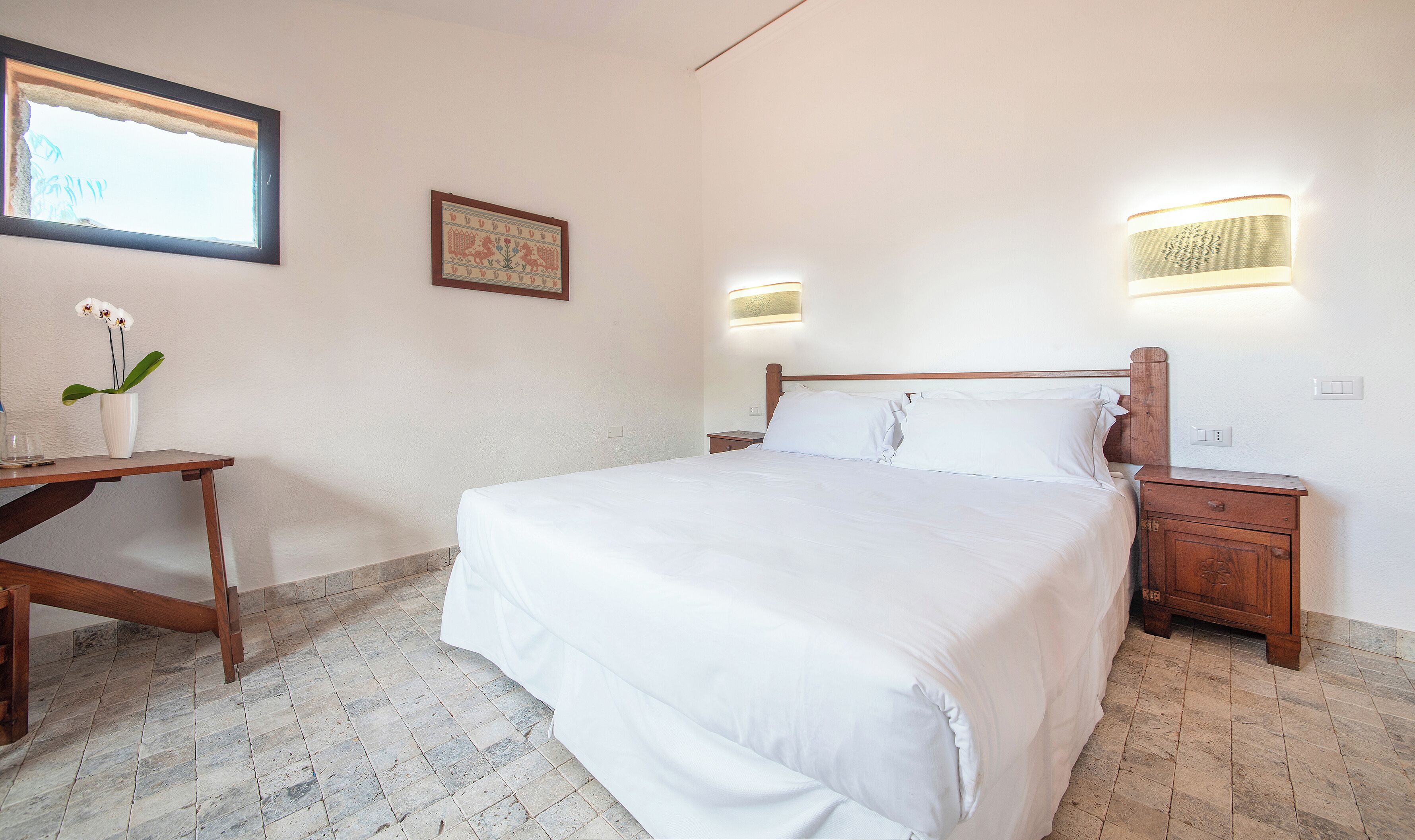 A neatly made bed with white linens, a bedside table with a vase of flowers, and a wall-mounted light fixture.
