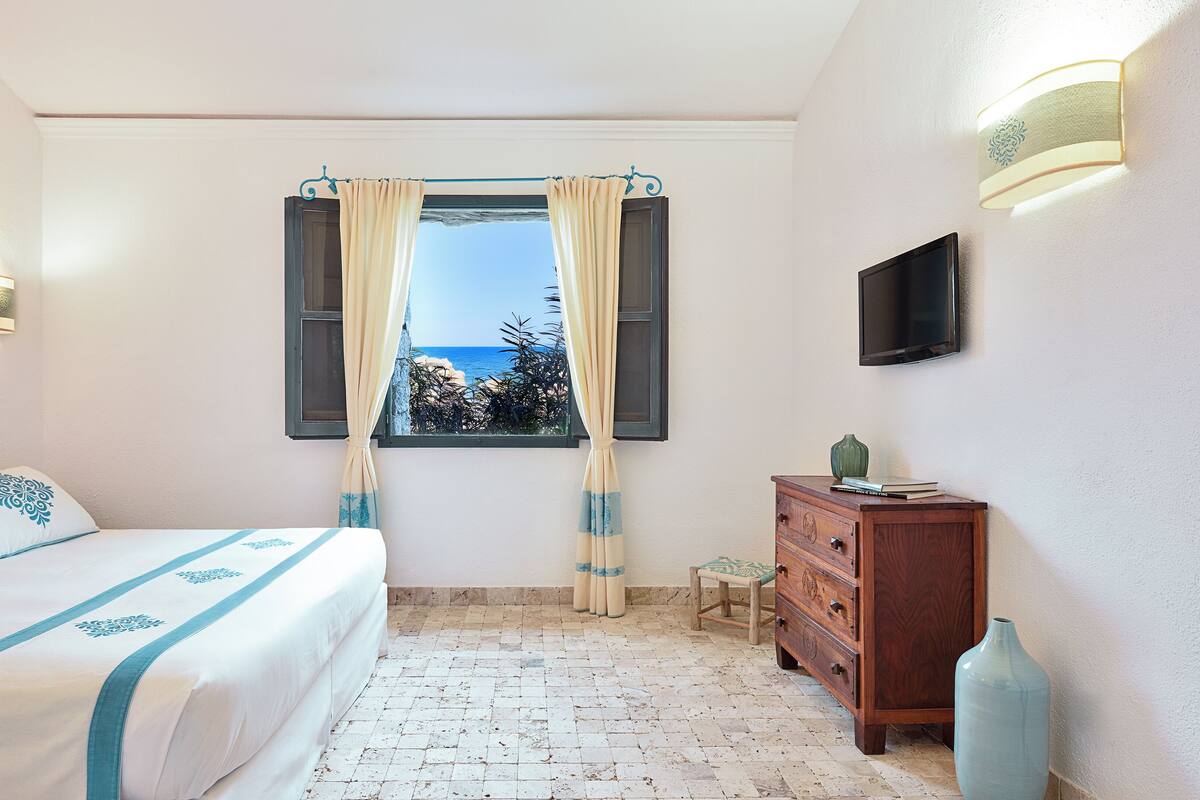 A bedroom with a bed, a dresser, a small table, a TV, and a window with curtains.