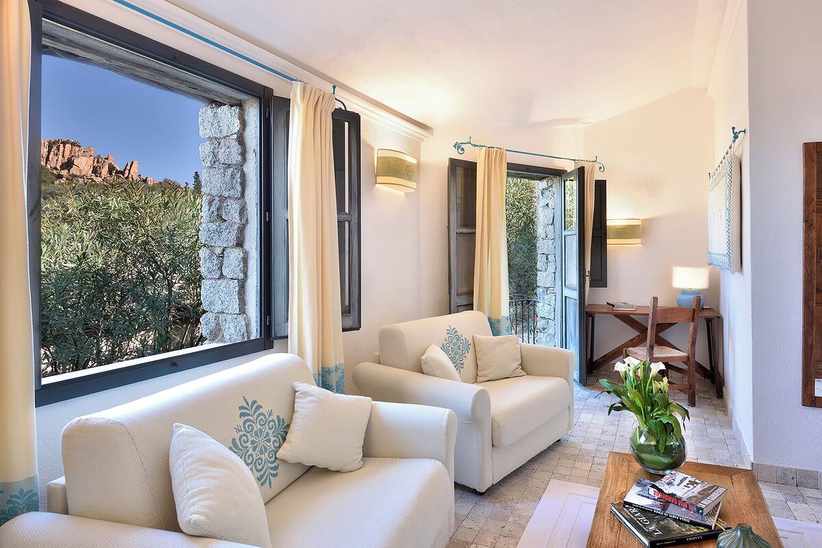 A spacious living area with a large window, two sofas, a wooden table, and a small desk with a lamp.