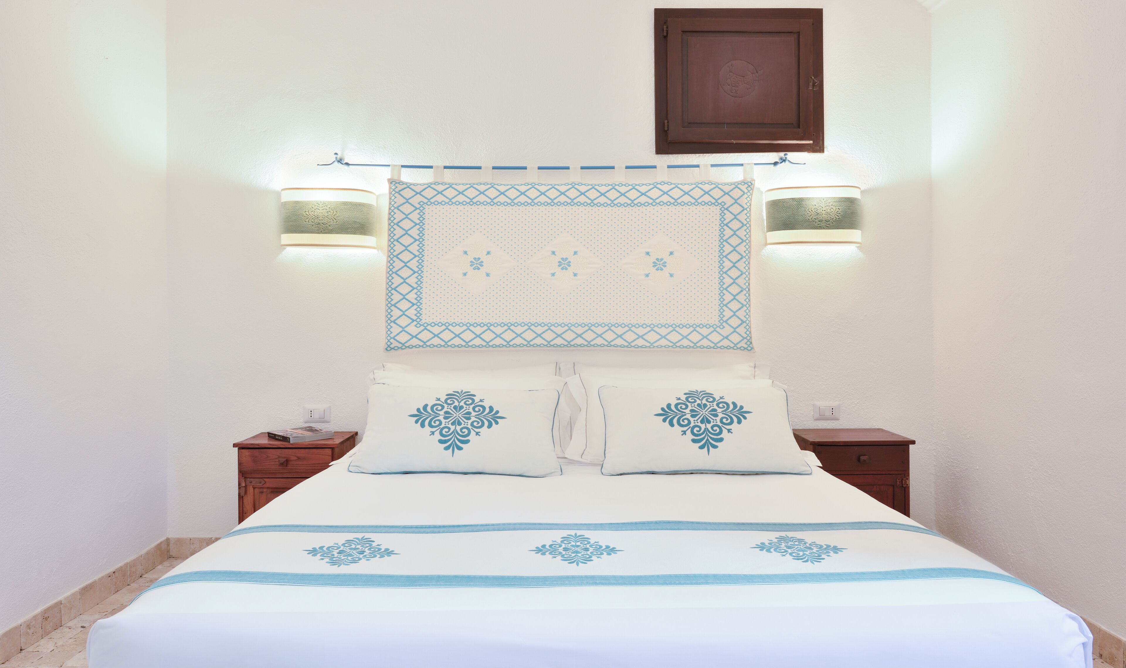 A neatly made bed with a white and blue bedspread, two pillows with blue patterns, and two wall-mounted lamps.