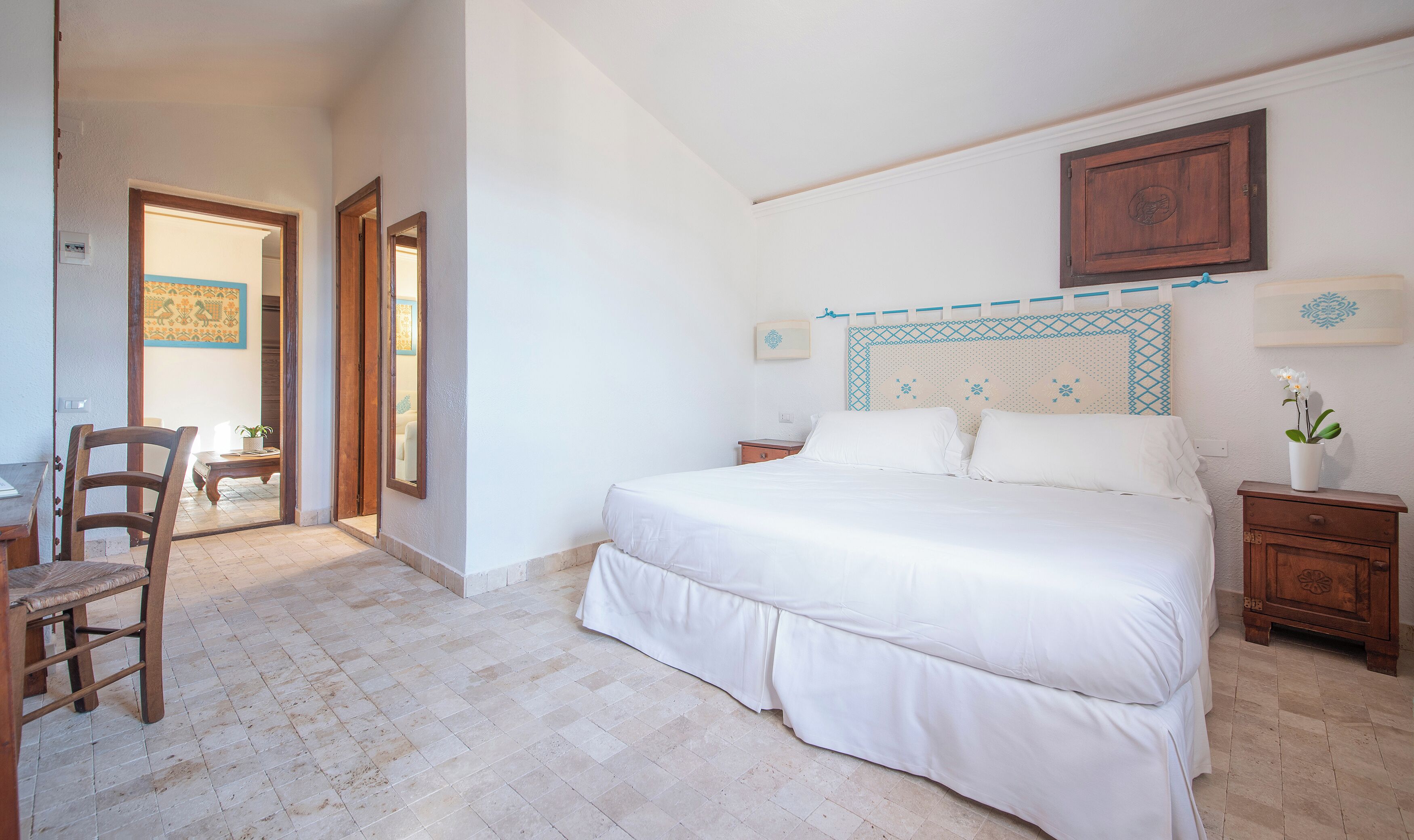 Ein Schlafzimmer mit einem großen Bett, einem Holfnachtkästchen, einem Stuhl und einer Tür zu einem anderen Raum.