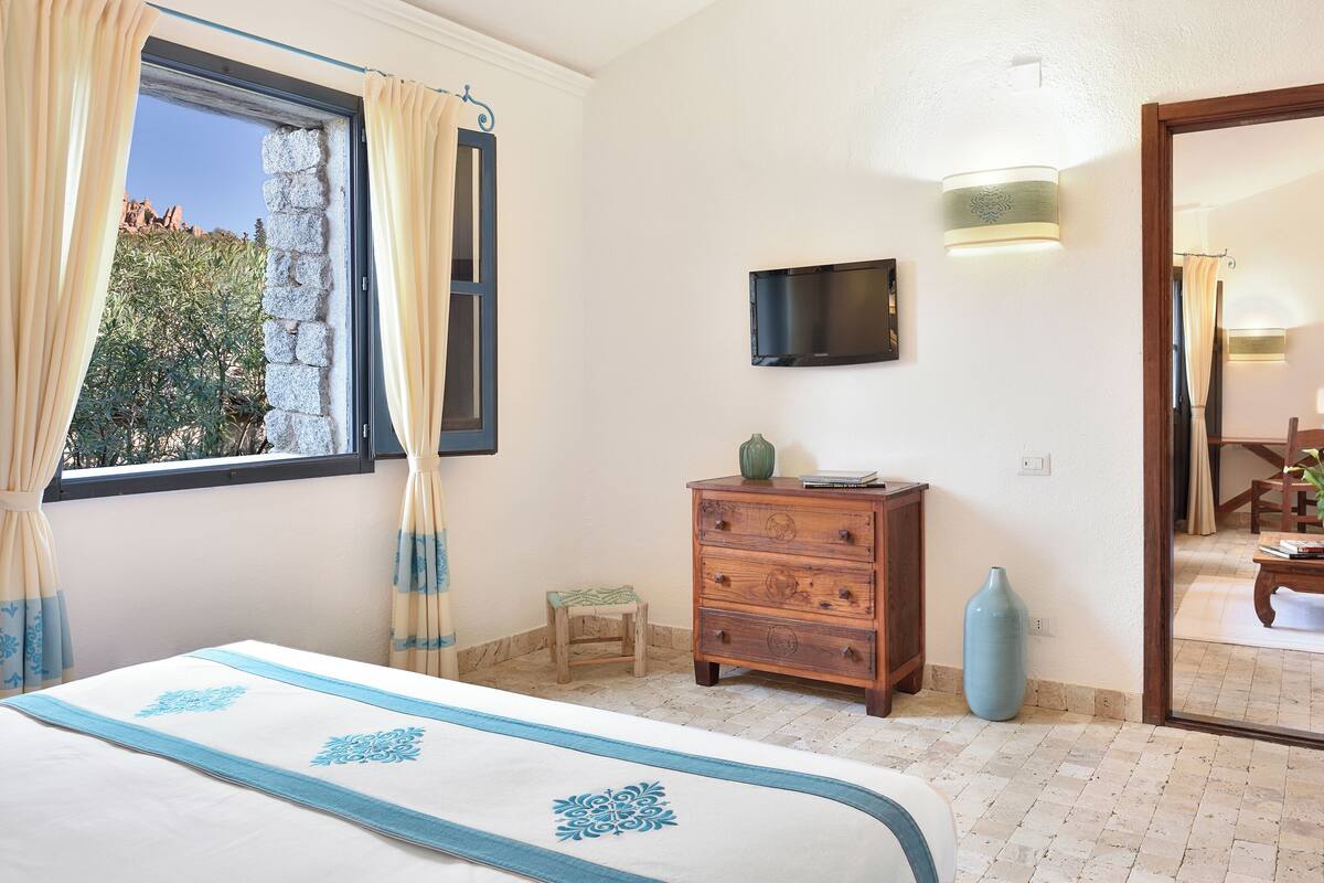 A bedroom with a bed, a wooden dresser, a television, and a window with curtains.